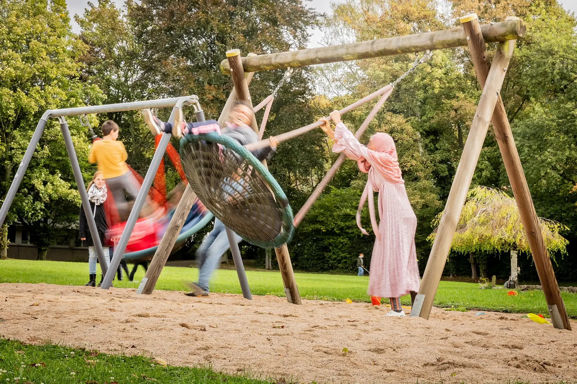 Zwei Nestschaukel auf dem Schul-Außengelände. Der Untergrund ist mit feinem Sand bedeckt. In beiden Schaukelkörben sitzen Schüler*innen, die von jeweils eine Lehrkraft angeschaukelt werden. Im Hintergrund sieht man frische Bäume und Graswiesen.