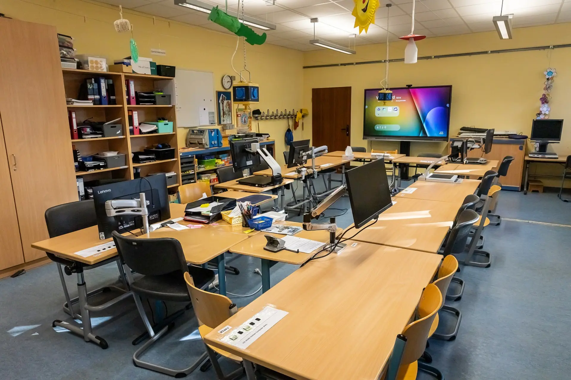 Der Klassenraum ist mit individuell angepassten Schülerarbeitsplätzen ausgestattet, die jeweils über Monitore und spezielle Hilfsmittel verfügen. Im Hintergrund hängt ein großer interaktiver Bildschirm an der Wand. Direkt daneben ist die Zimmertür. An der linken Wand befindet sich ein offenes Regal mit Ordnern und Lernmaterialien und einer Hakenleiste für die Jacken der Schüler*innen. Die Wände sind in Farbe gestrichen. An den Wänden und von der Decke hängen einige Dekorationen angebracht, was eine freundliche Lernatmosphäre schafft.
