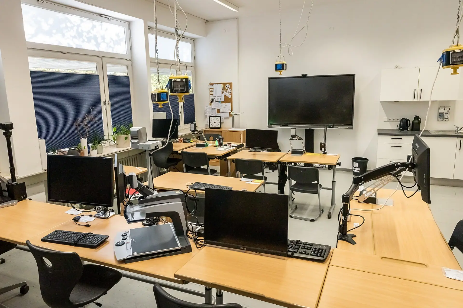 In diesem Klassenraum sind mehrere individuell angepasste Arbeitsplätze mit großen Bildschirmen und spezieller Hardware wie Zeichentabletts auf hölzernen Tischen verteilt. Jeder Platz verfügt über verstellbare Monitorarme und zusätzliche Beleuchtung von der Decke. Im Hintergrund befindet sich eine Küchenzeile mit Schränken und ein großer Flachbildschirm an der Wand. Große Fenster auf der linken Seite lassen viel Tageslicht herein.