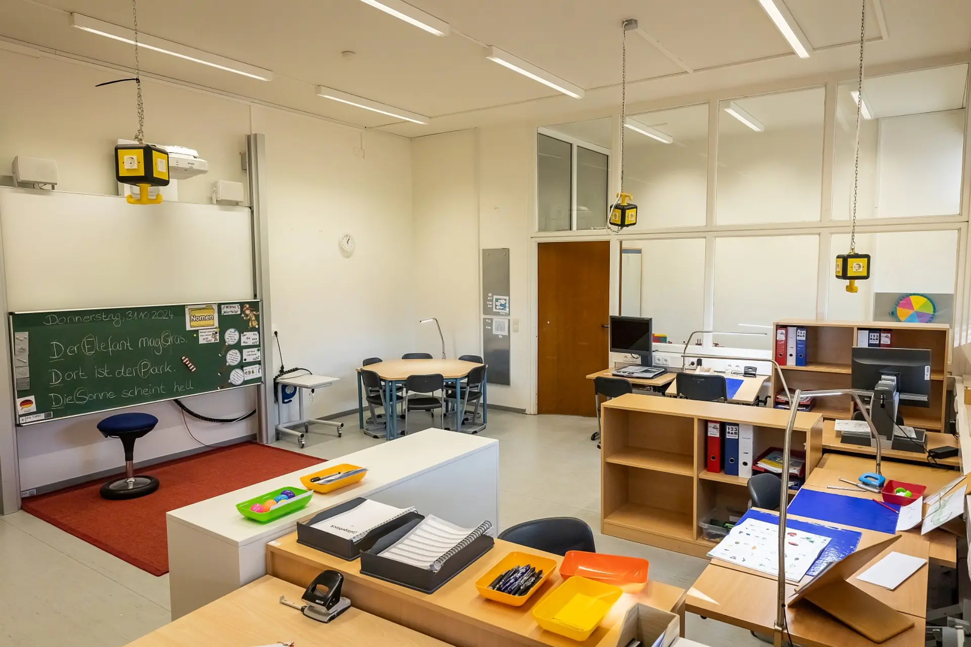 In diesem geräumigen Klassenraum für die Primarstufe befindet sich links eine Schultafel mit handschriftlichen Notizen und eine weiße Projektionsfläche dahinter, sodass man beides benutzen kann. Im Vordergrund sind Tische mit Lernmaterialien, Stiften und zusätzlicher Ausleuchtung an jedem Tisch zu sehen. Im hinteren Bereich gibt es sowohl einen Gruppentisch mit Stühlen für sechs Personen, als auch individuelle Arbeitsplätze mit Computern und Regalen mit Ordnern. Die Decke ist mit Neonröhren-Lampen ausgestattet, an paar Stellen hängen Steckdosen-Würfel mit Hacken zum Aufhängen und Anschalten von zusätzlichen Geräten. Die Wand rechts, wo sich auch die Zimmertür befindet, ist komplett aus Glas, sodass man dadurch zum Flur hinausschauen kann.