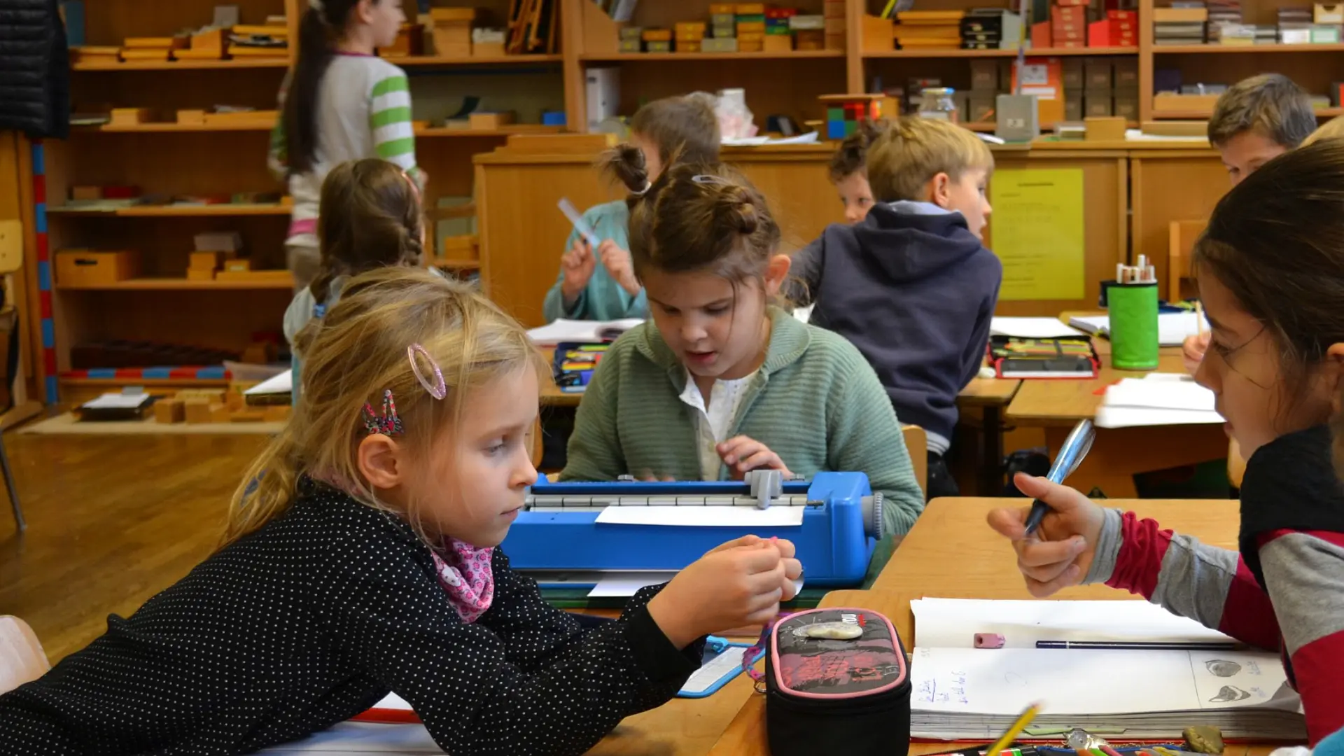 Ein Klassenzimmer mit Holzschreibtischen und vielen Schulkindern mit und ohne Sehbeeinträchtigung, die alleine oder in Gruppen an ihren Schulaufgaben arbeiten. 