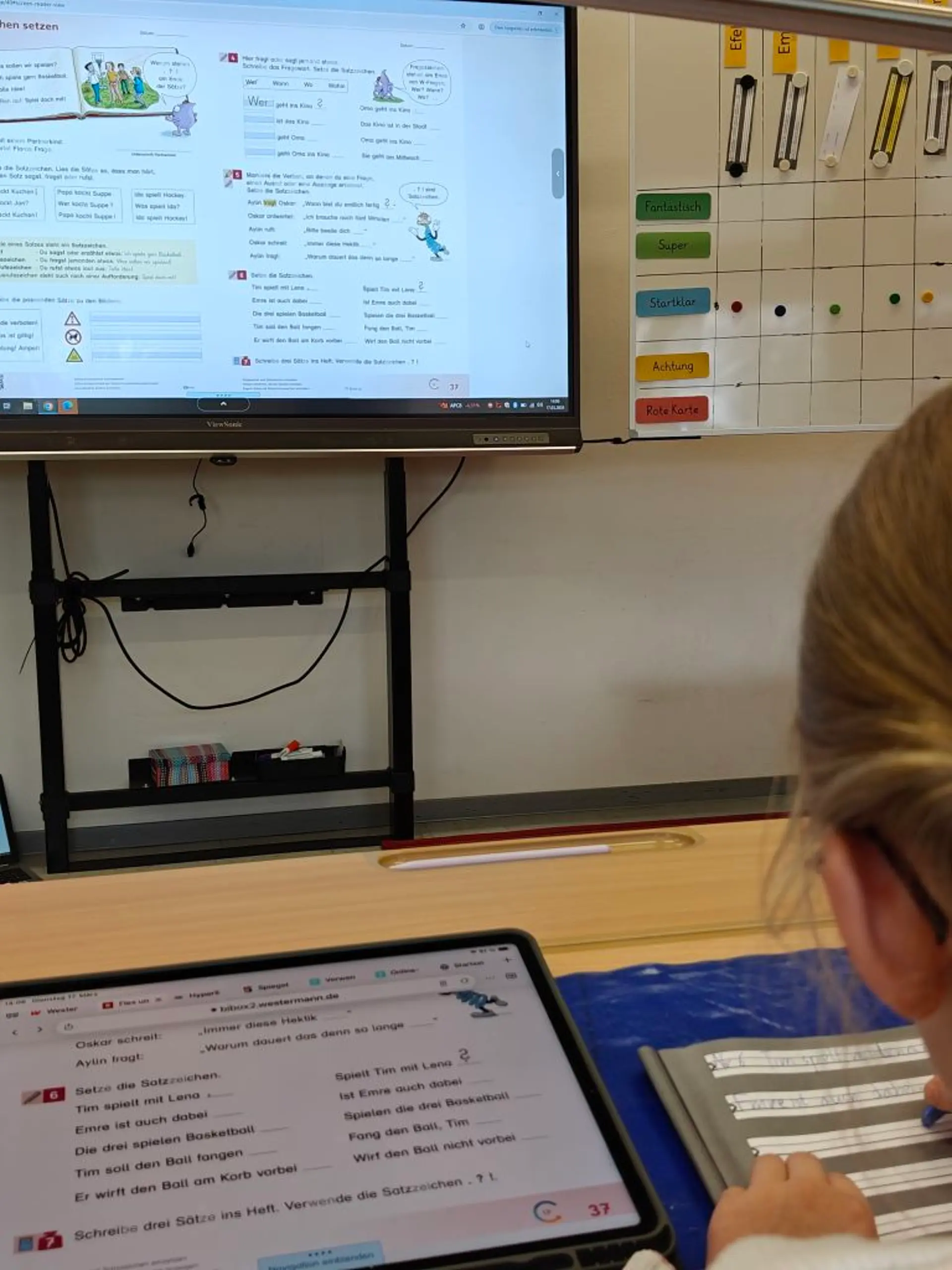 Blick über die Schulter einer Schülerin, die am IPad liest und auf speziellen Lineaturen schreibt