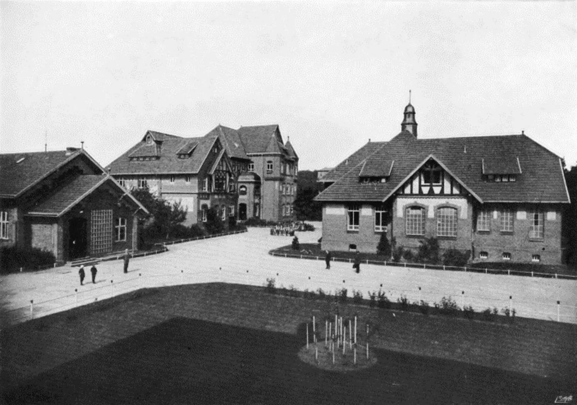 Ein altes, monochromes Foto zeigt eine erweiterte Ansicht der Blindenanstalt, die mehrere Gebäude auf einem weitläufigen Gelände umfasst. Im Vordergrund befindet sich eine große Freifläche, die teils begrünt, teils gepflastert ist, mit einigen kleinen Strukturen. Im Mittelgrund dominieren zwei größere Gebäude: rechts ein markantes Gebäude mit Fachwerkdetails und einem turmartigen Aufsatz auf dem Dach, links ein weiteres Gebäude mit einer großen Fensterfront. Im Hintergrund ist ein größeres, komplexes Gebäude mit mehreren Giebeln und Erkern zu sehen. Auf dem Gelände sind mehrere kleine, menschenähnliche Figuren zu erkennen, die sich bewegen.