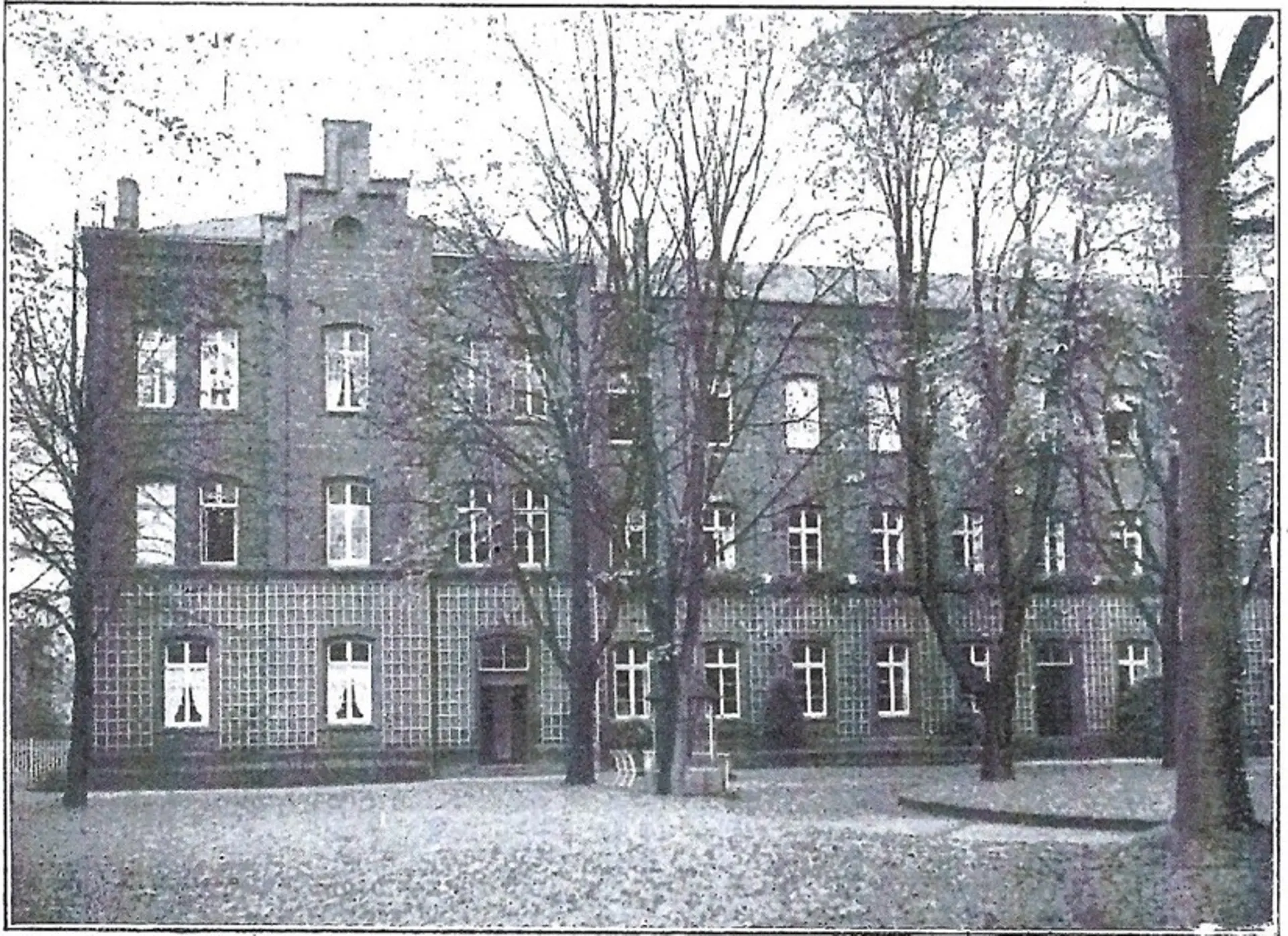 Ein monochromes Foto zeigt ein großes, mehrstöckiges Gebäude, das Teil der erweiterten Blindenanstalt ist. Die Fassade des Gebäudes ist aus dunklen Ziegeln und weist zahlreiche Fenster auf, die in Reihen angeordnet sind. Im unteren Bereich des Gebäudes ist eine auffällige, Gitter-artige Textur zu erkennen. Vor dem Gebäude befindet sich ein offener Bereich, der teils als Rasenfläche, teils als unbefestigter Boden erscheint. Mehrere hohe Bäume mit kahlen Ästen stehen im Vordergrund und verdecken teilweise die Ansicht des Gebäudes, was auf den Spätherbst oder Winter hindeutet.