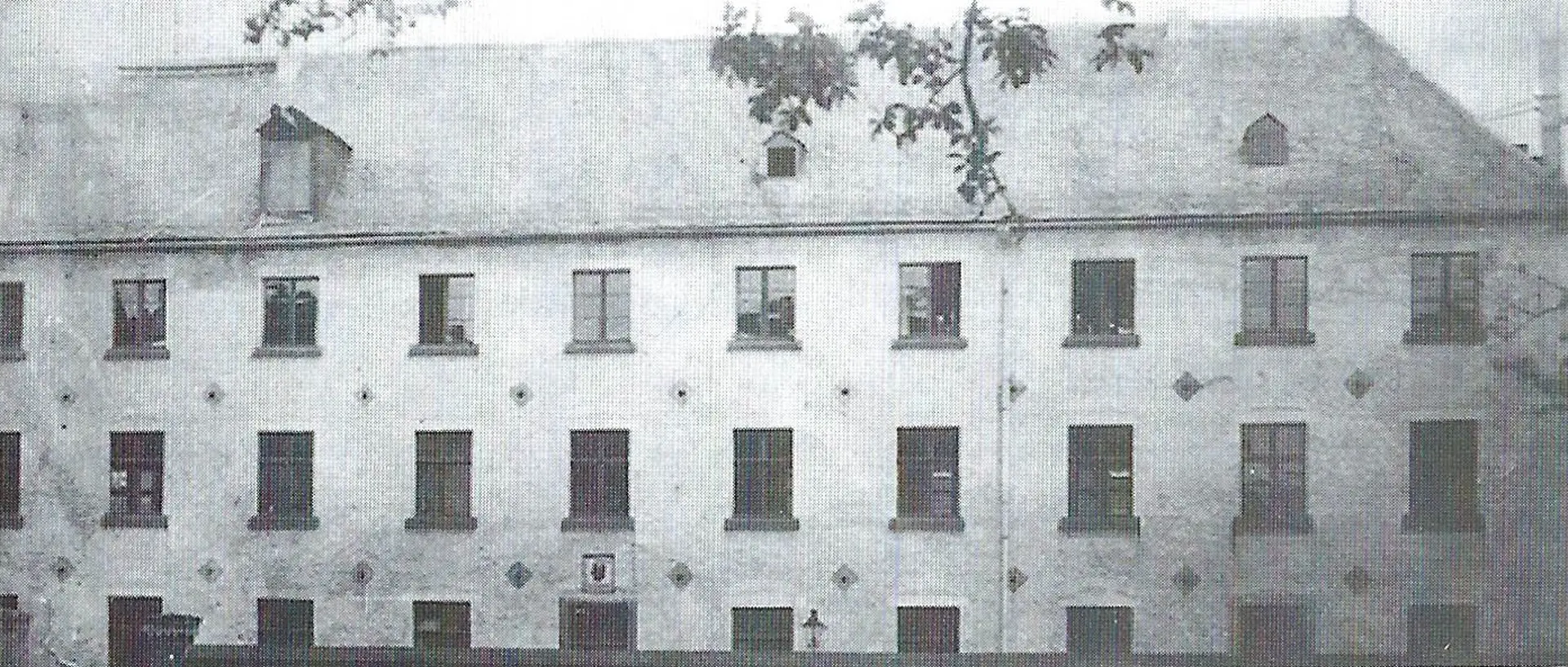 Ein monochromes, historisches Foto zeigt das ehemalige Jesuitenkloster, das um 1910 als Blindenanstalt genutzt wurde. Das Gebäude ist ein mehrstöckiges, rechteckiges Bauwerk mit zahlreichen Fenstern, die in Reihen angeordnet sind. Das Dach ist steil und besitzt mehrere Dachgauben. Die Fassade weist eine schlichte Gestaltung auf, wobei im unteren Bereich des Gebäudes ein Wappen oder Emblem zu erkennen ist.