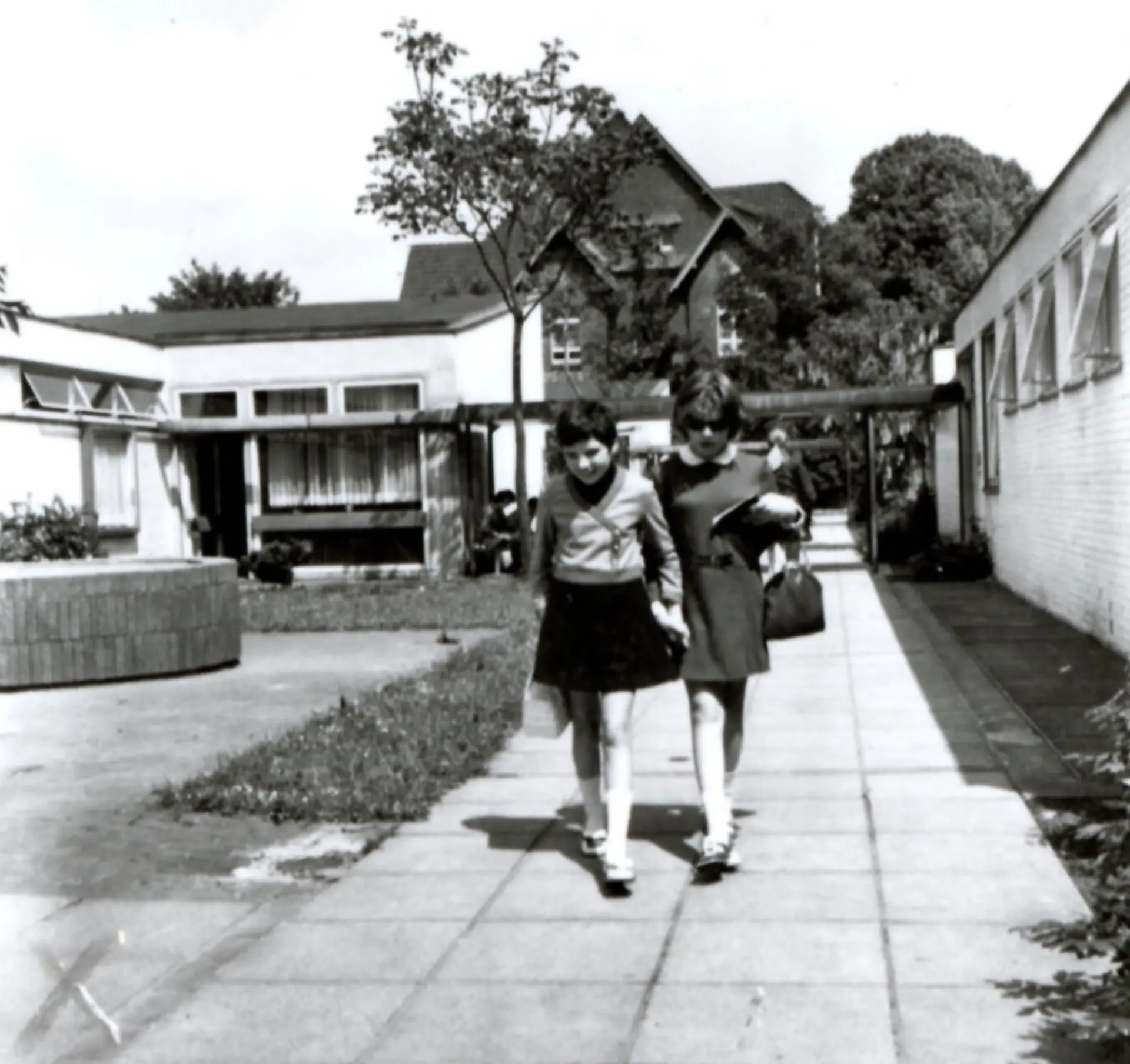 Eine Schwarzweiß-Aufnahme von zwei Schüler*innen, die auf dem Fußweg zwischen den Gebäuden auf dem Schulhof laufen. Es wirkt so, als ob beide eine Sehbeeinträchtigung haben. Sie halten Hand, die eine Schülerin hat eine Sonnenbrille an, beide schauen zum Boden. Drum-herum sind frische Grasflächen sowie große Bäume mit üppigen Baumkronen zu sehen.