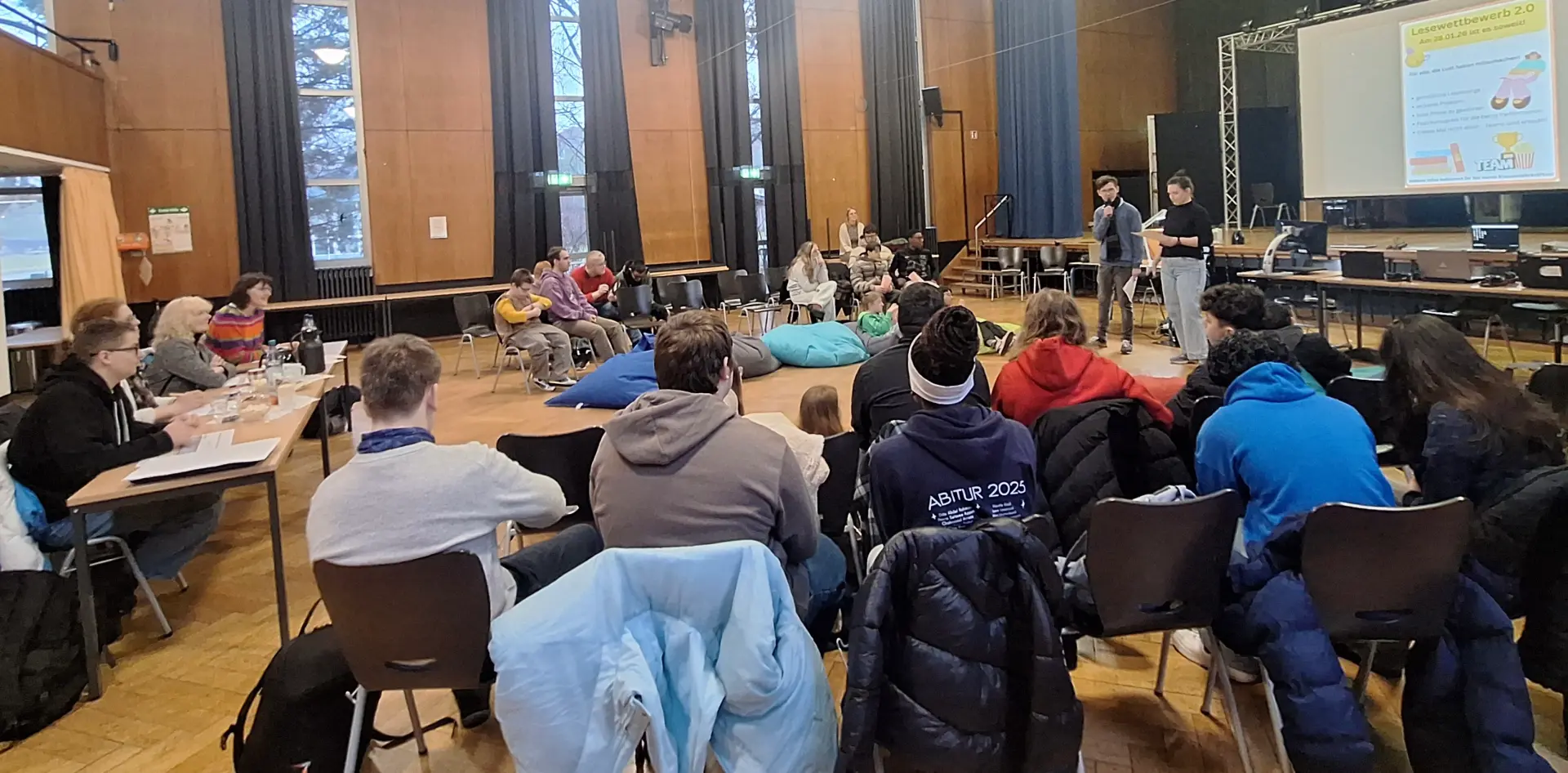 Schüler*innen und Jury sitzen im Halbkreis in der Aula. Vorne ist das Moderationsduo vor einer Leinwand zu sehen, auf die das Plakat vom Lesewettbewerb projiziert ist. Die Schüler*innen sitzen sowohl auf Stühlen als auch auf Sitzsäcken.