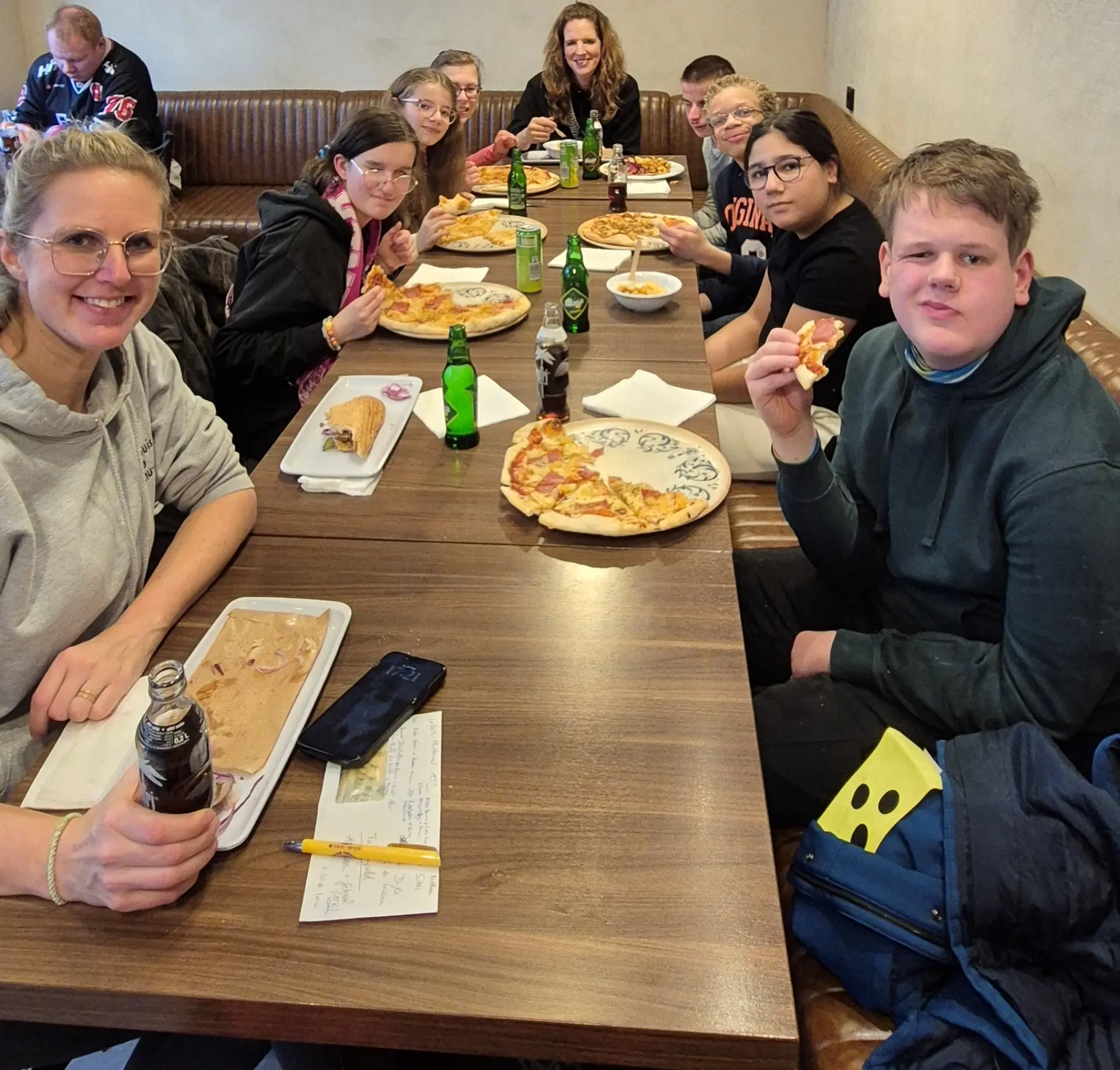 Die gesamte Gruppe sitzt an einem langen Tisch mit Pizza, Döner, Pommes und Getränken.