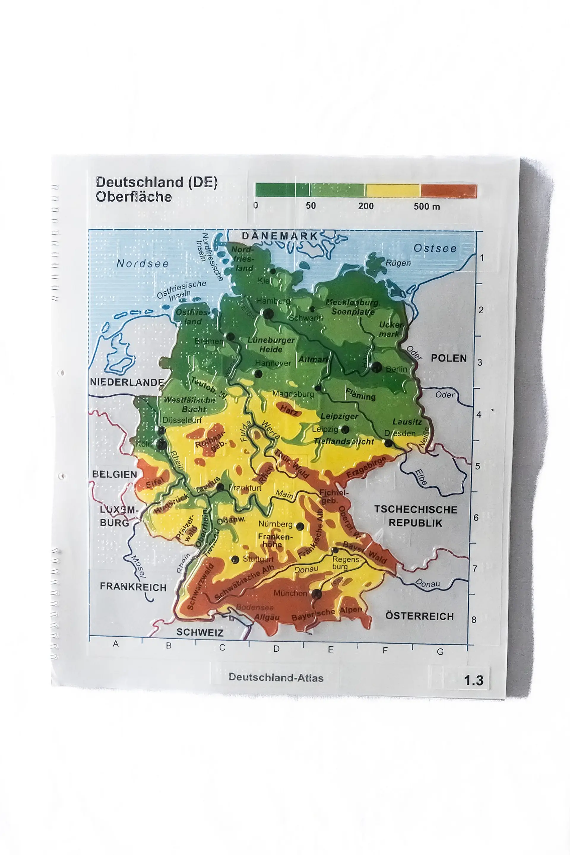 Taktile Karte von Deutschlands Oberfläche, das heißt, die Karte zeigt, wie hoch über dem Meeresspiegel einzelne Regionen gelegen sind. Die obere Hälfte des Landes ist vorwiegend flach mit bis zu 200 Metern über dem Meeresspiegel. Die untere Hälfte (vor allem in Baden-Württemberg und Bayern) wird es höher. Die Grenzen zu den benachbarten Ländern sind in Rot und dicker markiert, die großen Flüsse sind in Blau markiert. Die Hauptstädte der einzelnen Bundesländer sind mit einem dicken Punkt gekennzeichnet.. 