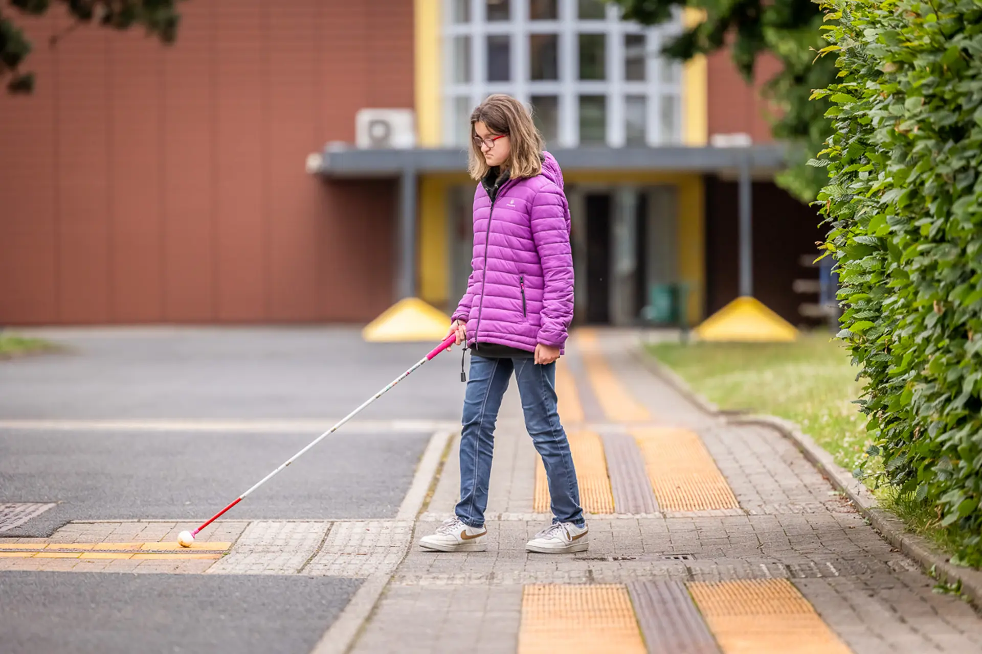 Eine Schülerin mit Blindheit läuft auf dem Weg zwischen den Schulgebäuden und orientiert sich mit einem Blindenstock anhand der Markierungen auf dem Boden. 