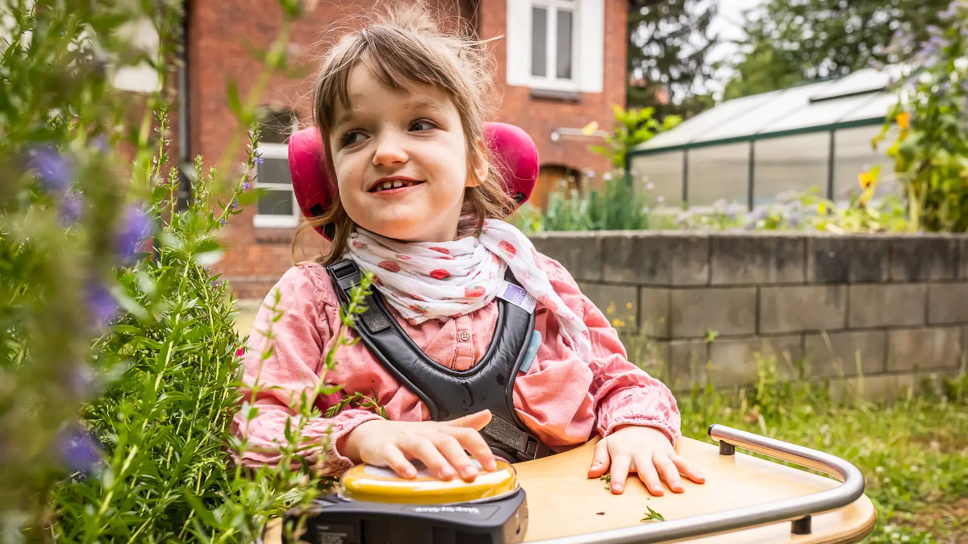 Eine Schülerin sitzt in einem speziellen Stuhl mit Nackenstütze und Tischplatte und drückt eine sprechende Taste, die vor ihr auf der Platte liegt. Die Schülerin befindet sich im Schulgarten und schaut seitlich zu einem blühenden Kräuterbusch neben ihr.