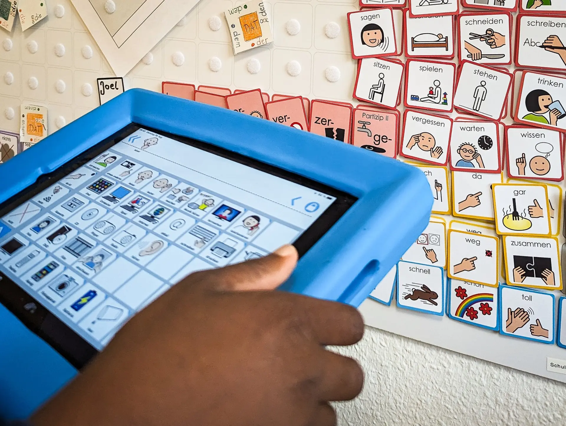 Die Hände eines Kindes, das ein Tablet hält, worauf verschiedene Metacom-Symbole zu sehen sind. Das Tablet ist auch zu einer Magnetwand gerichtet, woran viele einzelne Metacom-Symboltäfelchen zur Auswahl befestigt stehen. 