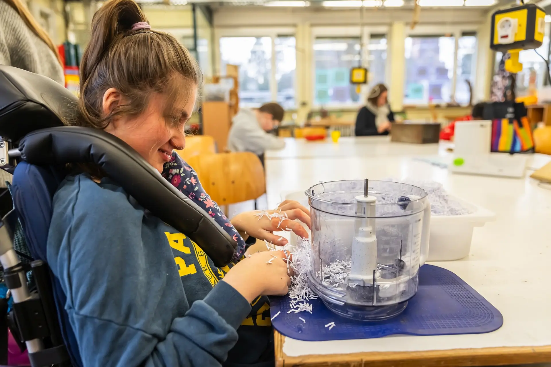 Eine Schülerin mit komplexer Beeinträchtigung sitzt im Rollstuhl vor einem Tisch im großen Schulraum. Vor ihr ist ein geöffneter Mixer, in den sie das geschredderte Papier reintun wird.