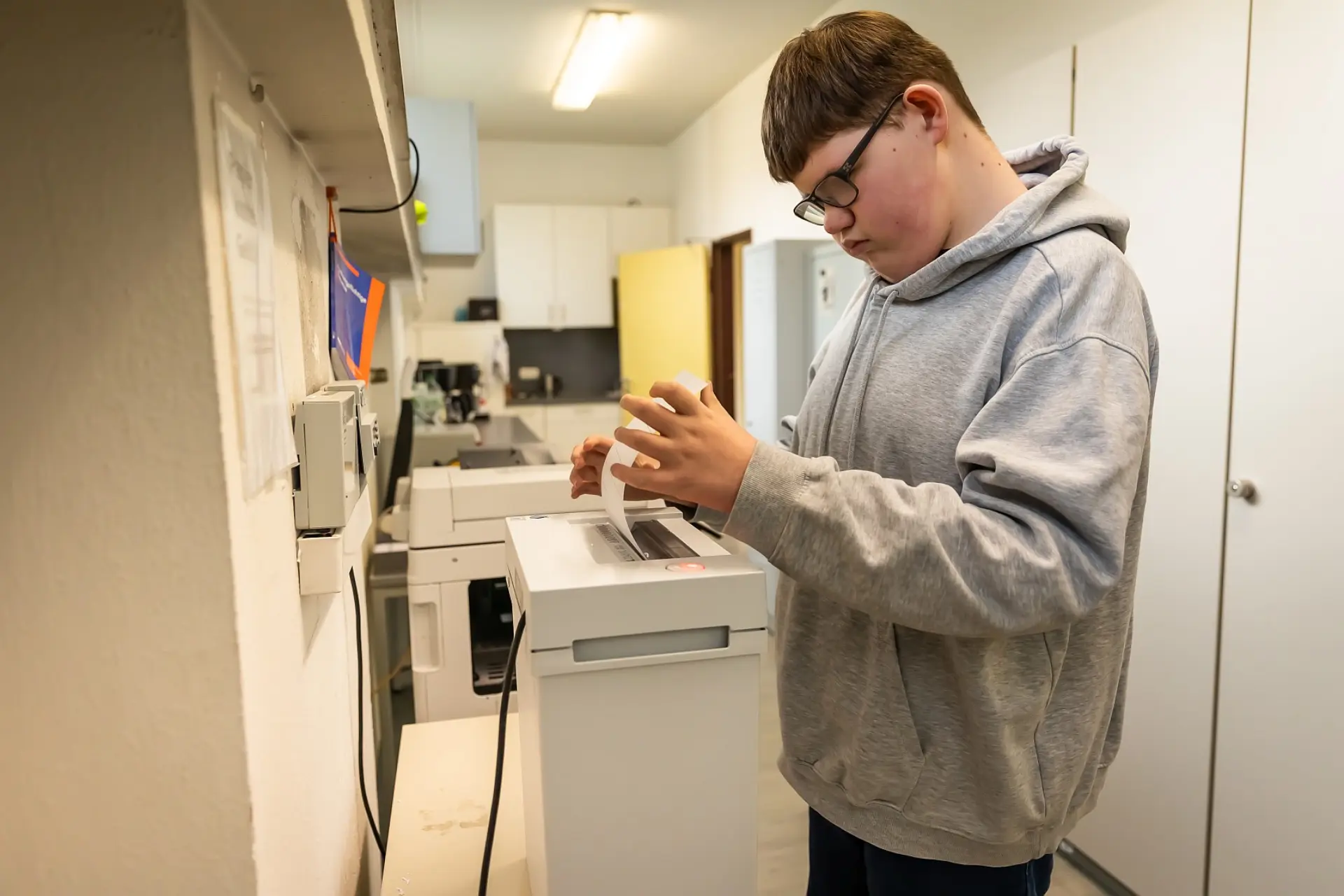 Ein Schüler steht vor einem Papierschredder in einem Schulbüroraum und steckt ein Blatt Papier in die Maschine ein. 