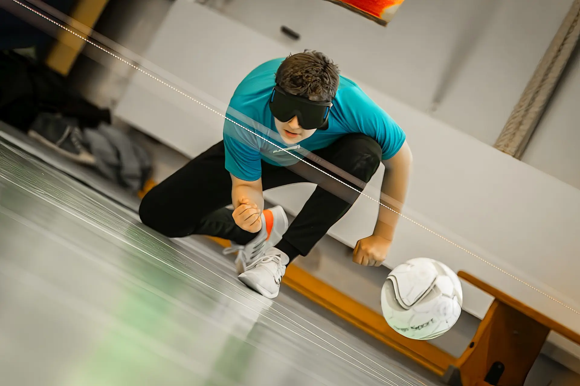 Schüler spielt Torball in einer Sporthalle. Er trägt eine große schwarze Brille, kniet auf dem Boden und hat gerade den Ball nach vorne geworfen. Der nach vorne fliegende Ball ist auch zu sehen. 