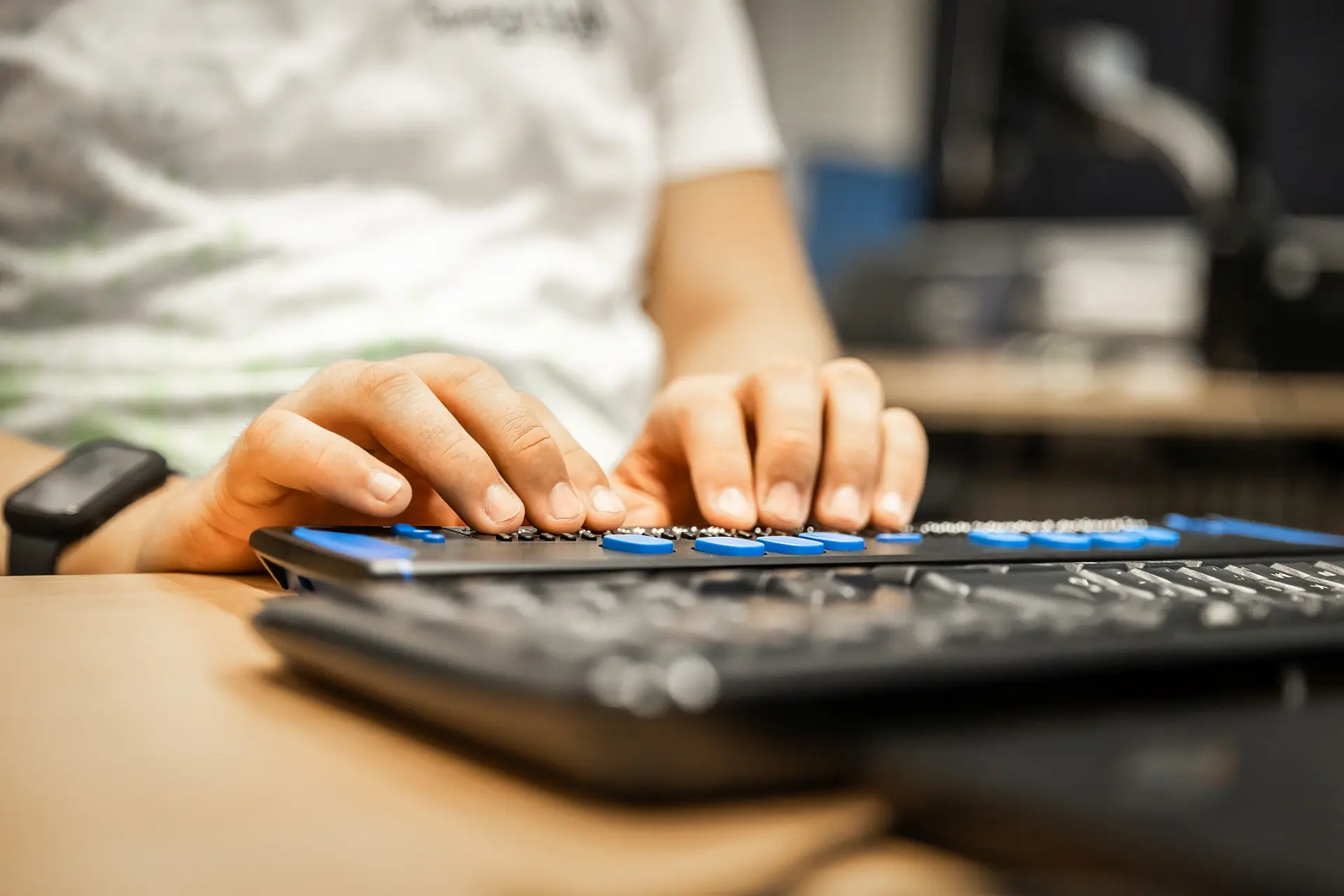 Die Hände eines Schülers, die an einer Tastatur für blinden Menschen tippen. Der Schüler befindet sich in einem Klassenraum