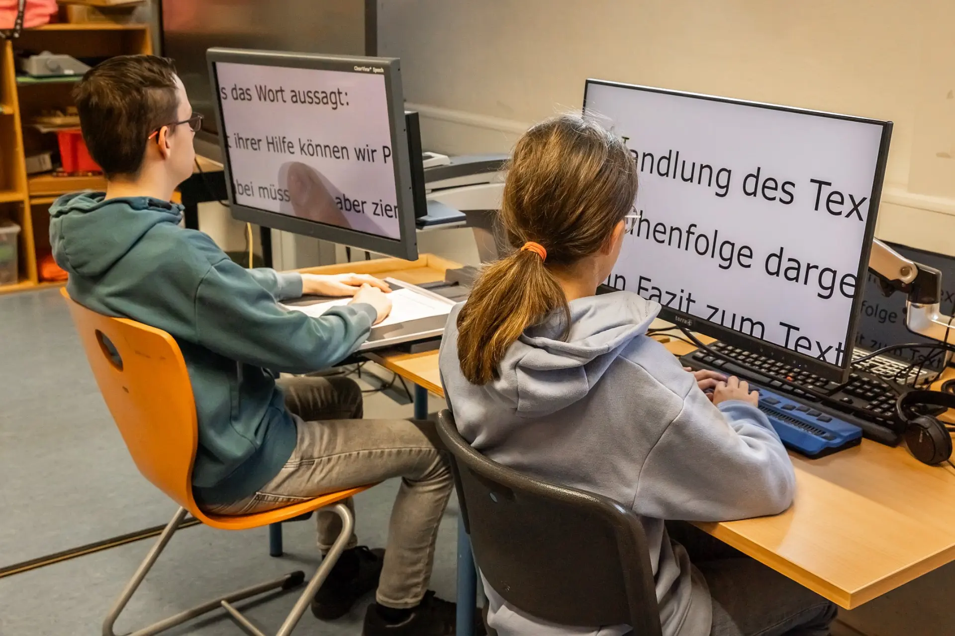 Zwei Schüler*innen sitzen am Schreibtisch in einem Schulraum. Vor jedem Jugendlichen sind jeweils ein Bildschirmlesegerät und eine Braillezeile, mit denen sie Texte lesen.