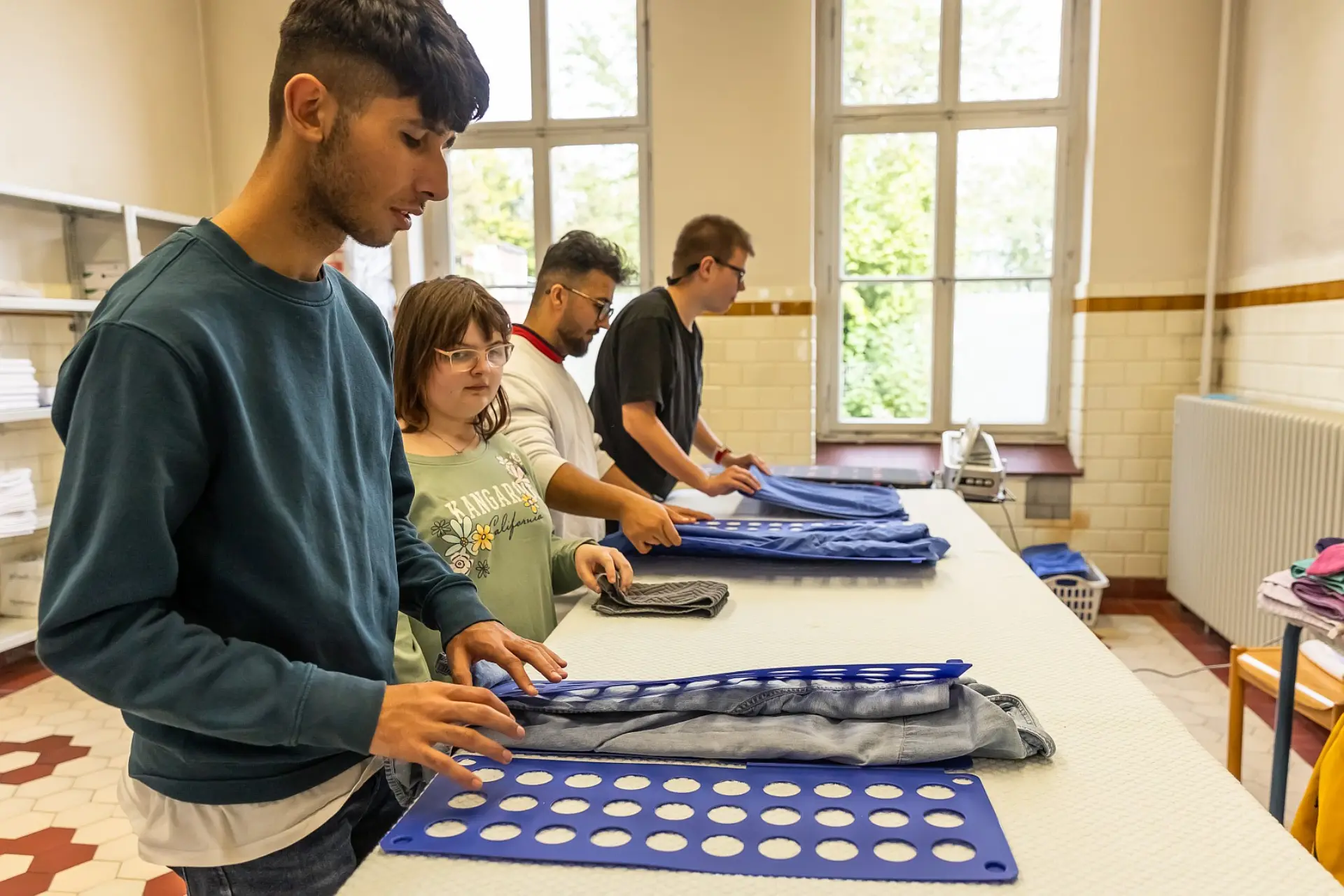 Vier Schüler*innen stehen vor einem langen Tisch und falten Wäsche mithilfe von Wäschefaltbrettern.