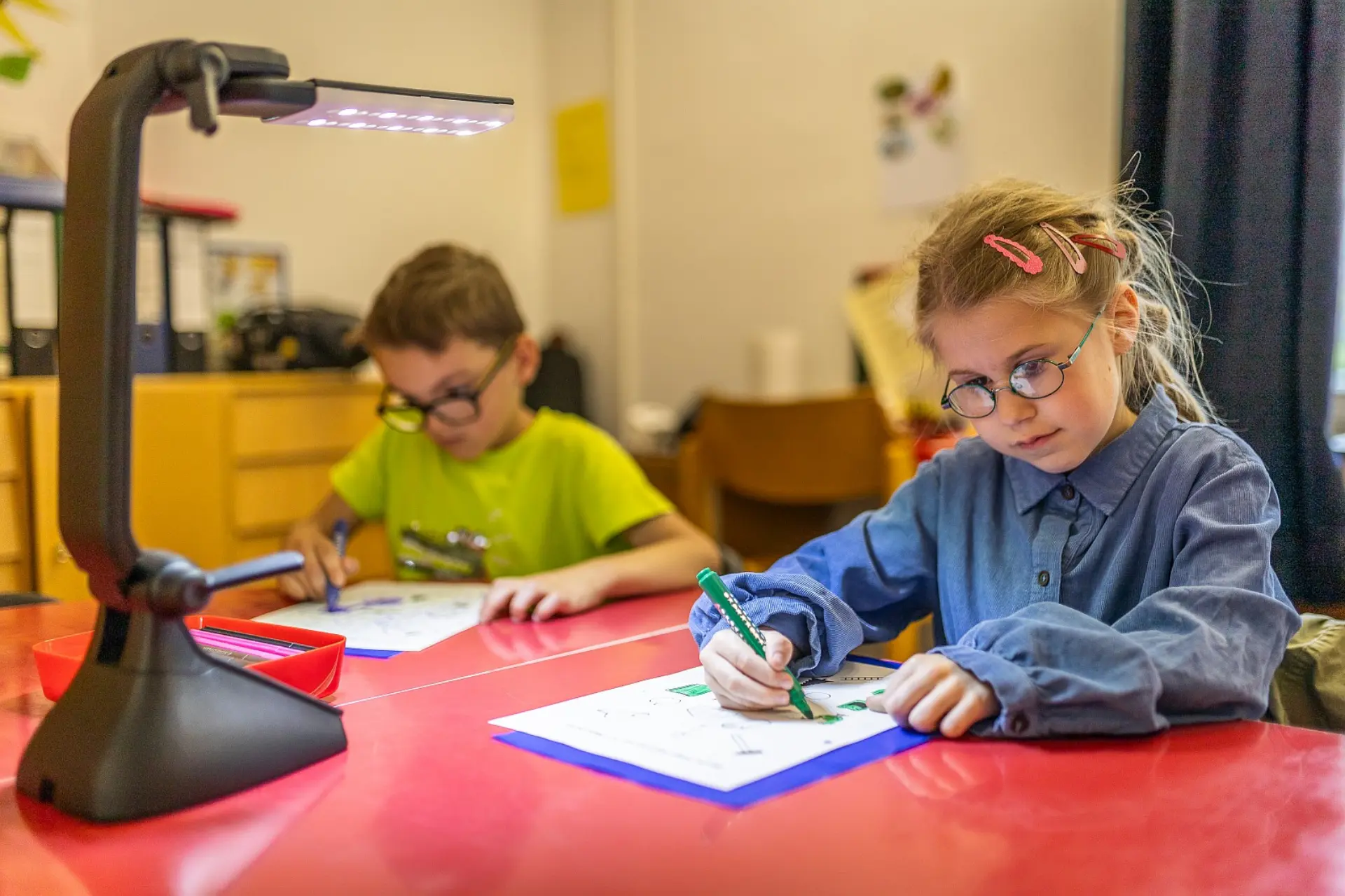 Zwei Kinder im Vorschulalter sitzen am Schreibtisch, leicht voneinander entfernt und füllen etwas in ihren Aufgabenheften aus. Ihre Arbeitsplätze sind mit Schreibtischlampen ausgeleuchtet und die Kinder sind in ihren Aufgaben vertieft.