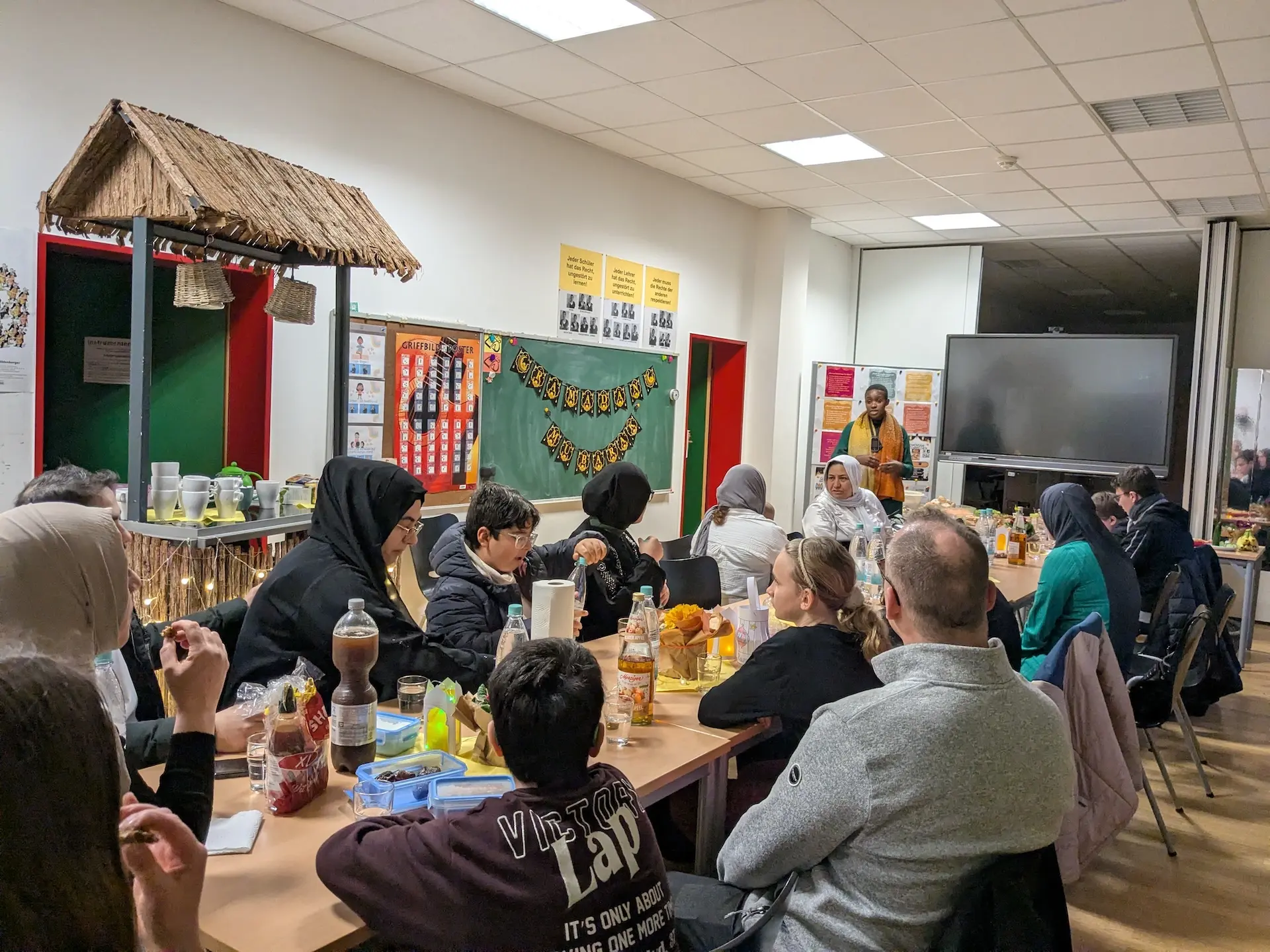 Familien sitzen an einem Tisch mit Deko und Getränken, eine Lehrerin erklärt etwas 
