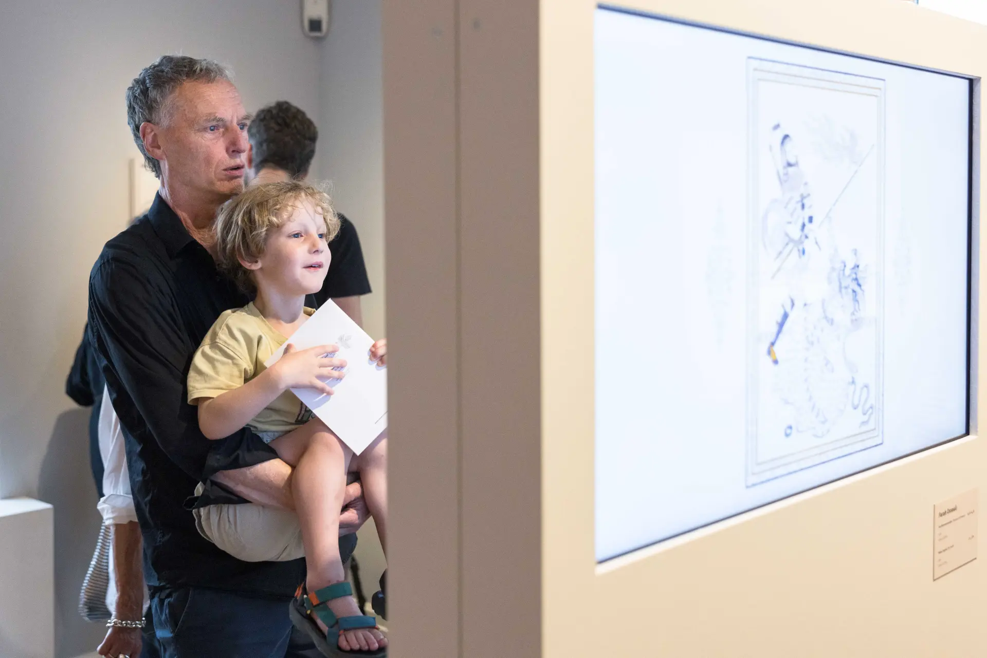 Ein Mann hält ein Kleinkind auf dem Arm, beide schauen sich auf einem Screen Kunstwerke an