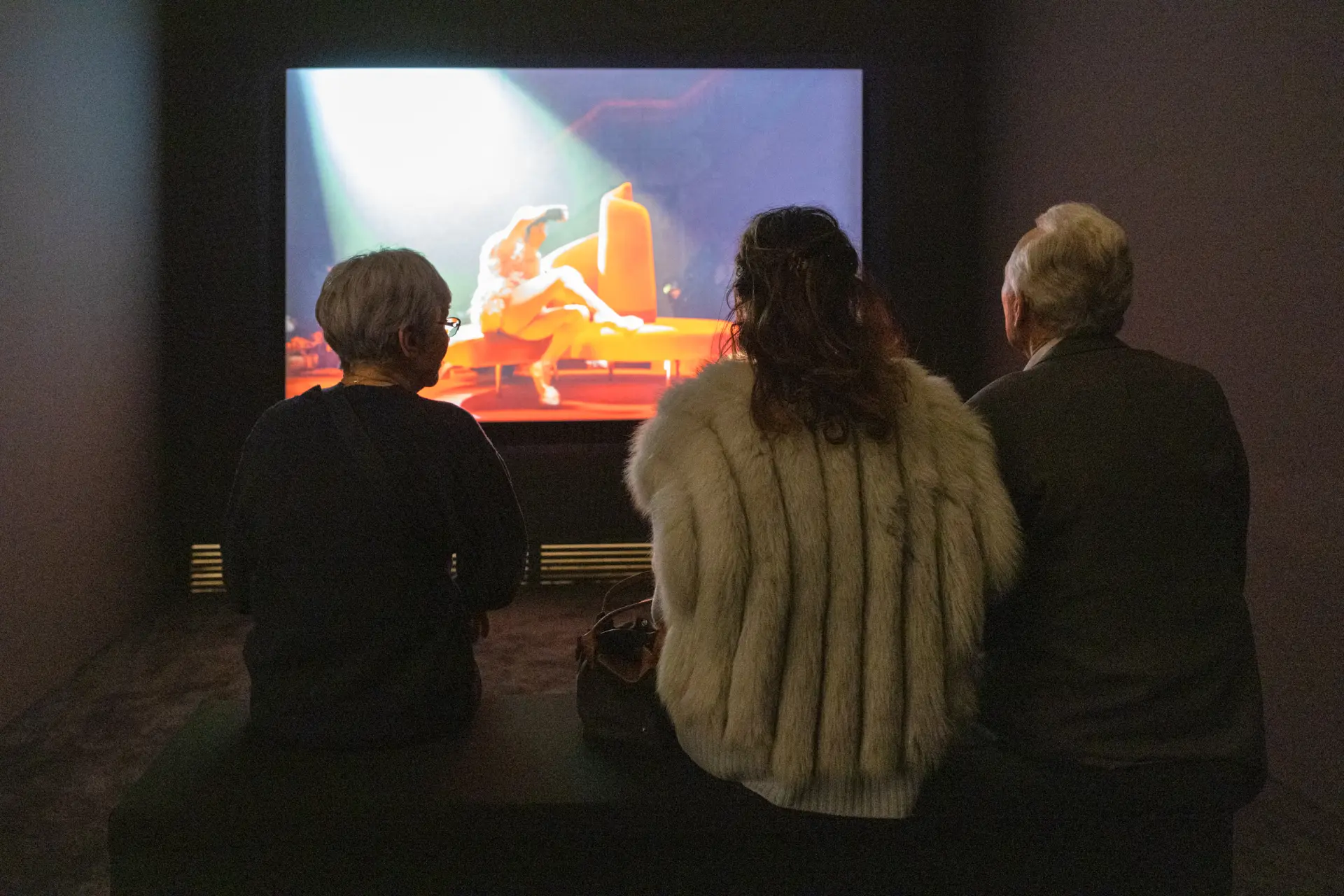 Eine Gruppe von drei Besucher*innen sitzt auf einem Sofa und schaut auf eine Video-Projektion