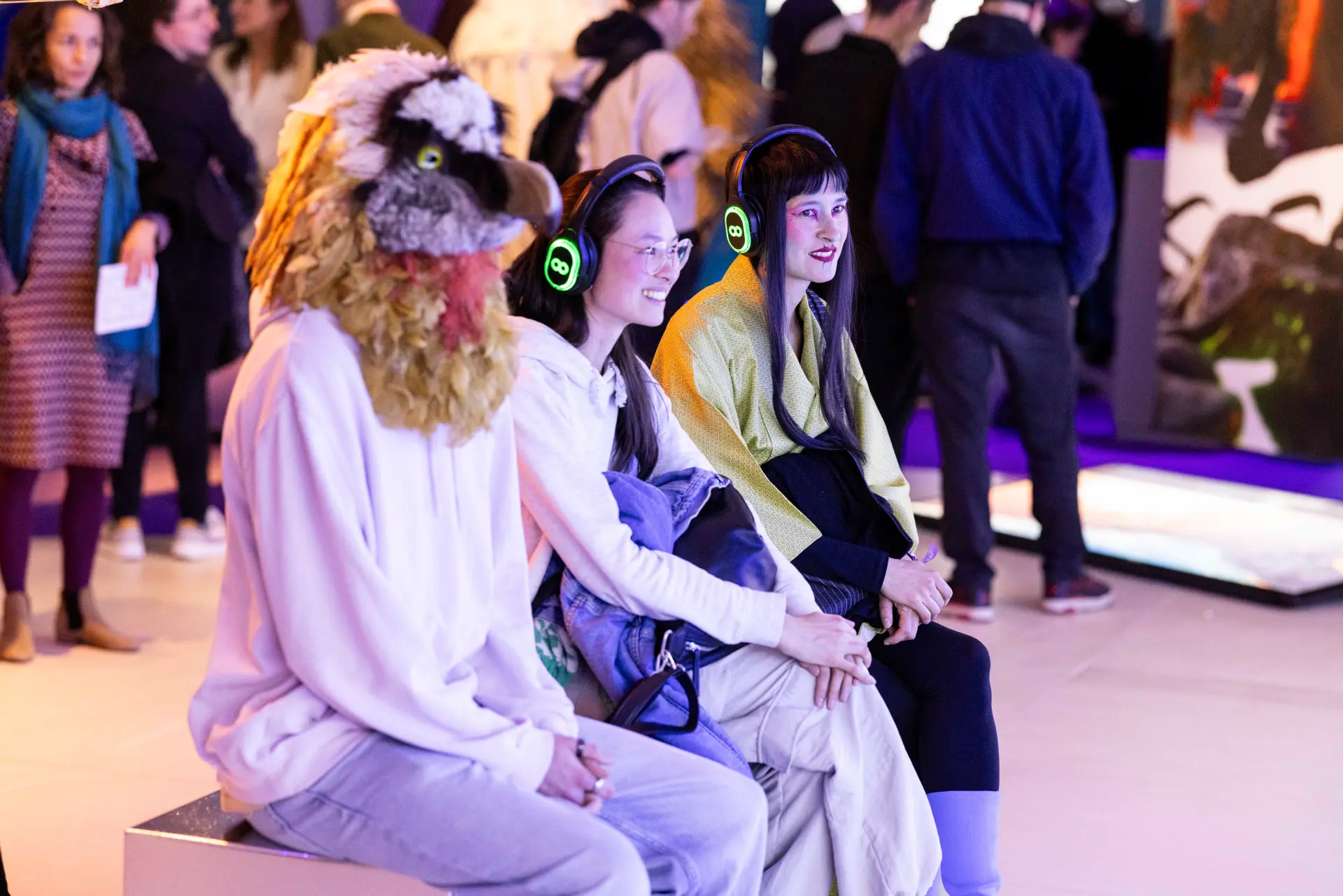 Besucherinnen sitzen mit Kopfhörern auf einer Bank, eine Person trägt eine Vogelmaske.