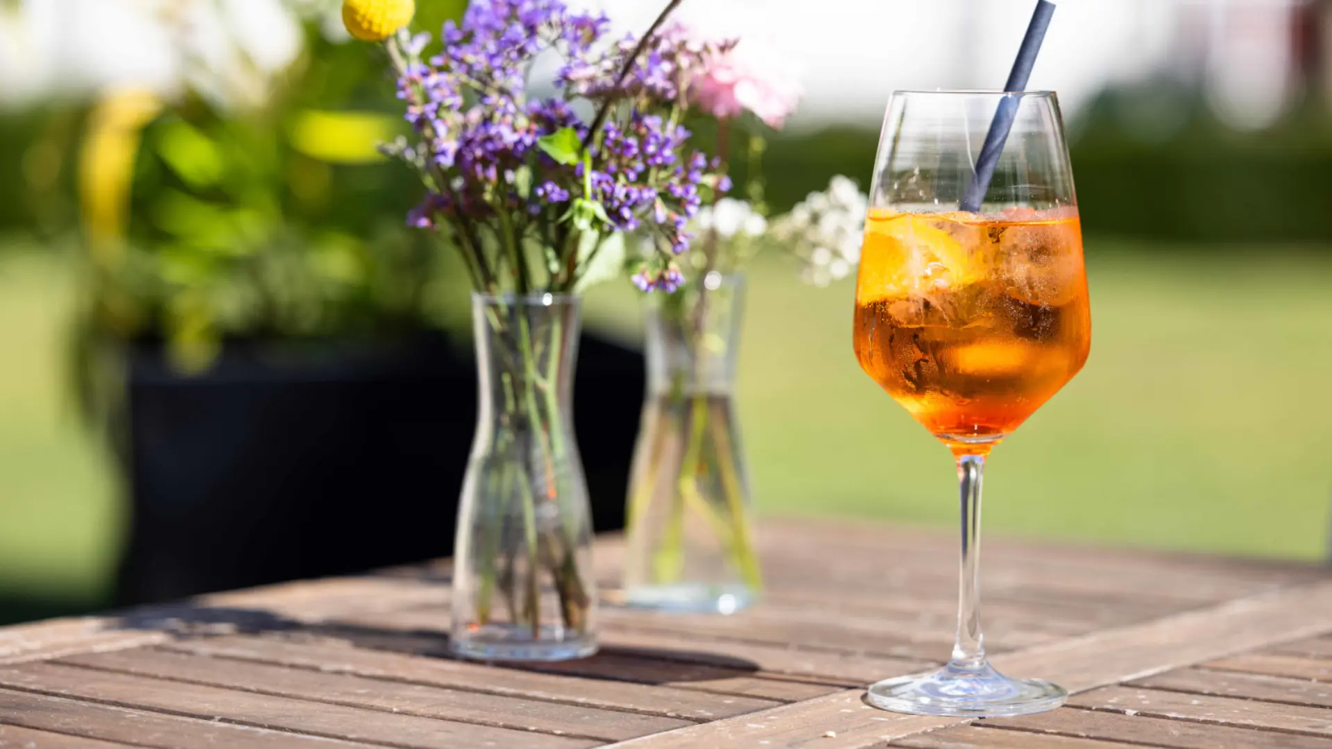 Ein Glas Aperol Spritz neben Vasen mit Blumen auf einem Tisch im Freien