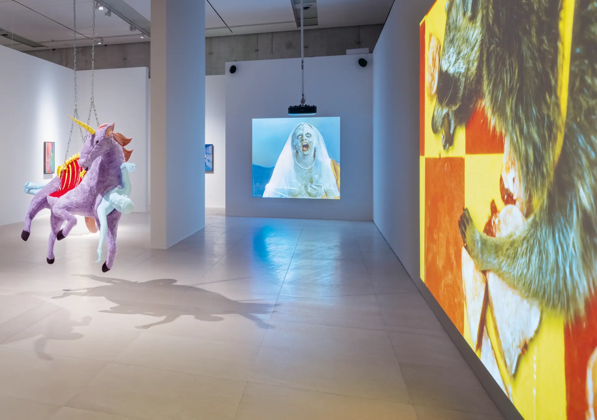 Ausstellungsansicht mit einer hängenden Einhornskulptur links und Videoinstallationen