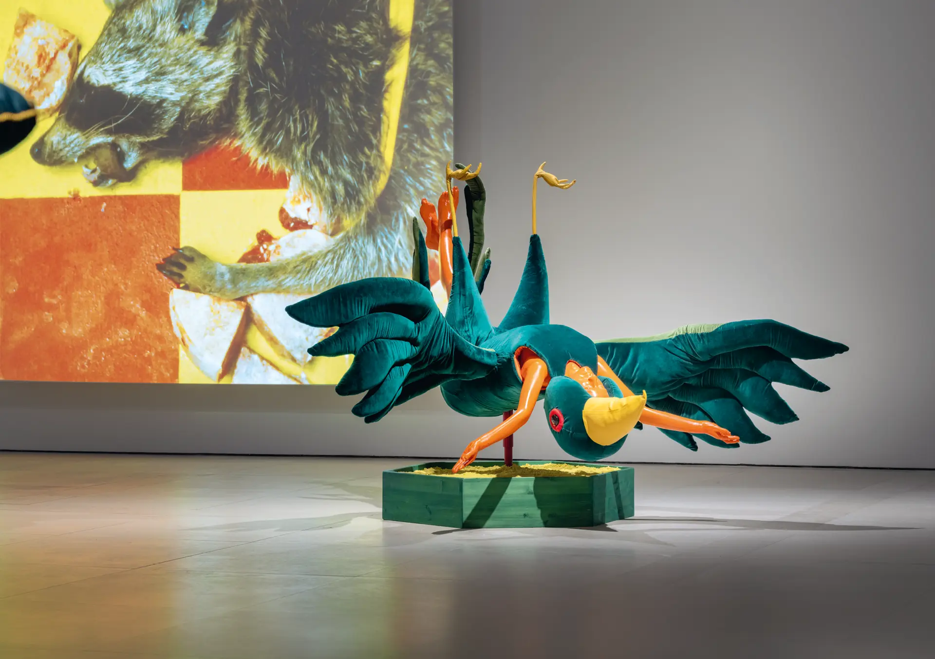 Ausstellungsansicht mit einer bunten textilen Vogelskulptur und einer Videoinstallation im Hintergrund