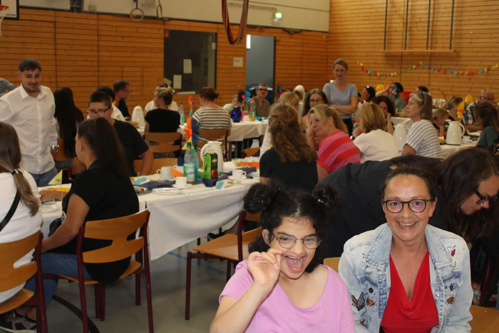 Blick in die Turnhalle. Es wird gemeinsam gefrühstückt. Im Vordegrund strahlen zwei Frauen in die Kamera