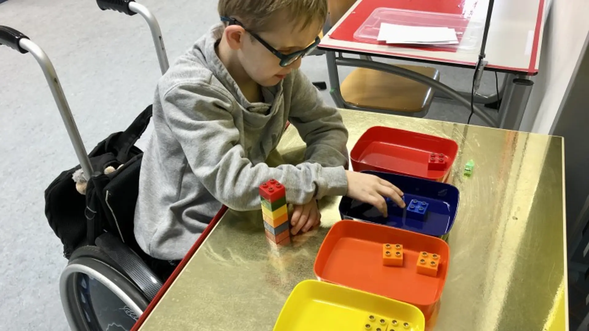 Ein Junge im Rollstuhl und mit einer starken Brille sortiert Lego-Duplo-Steine in verschiedenfarbige Plastikschalen
