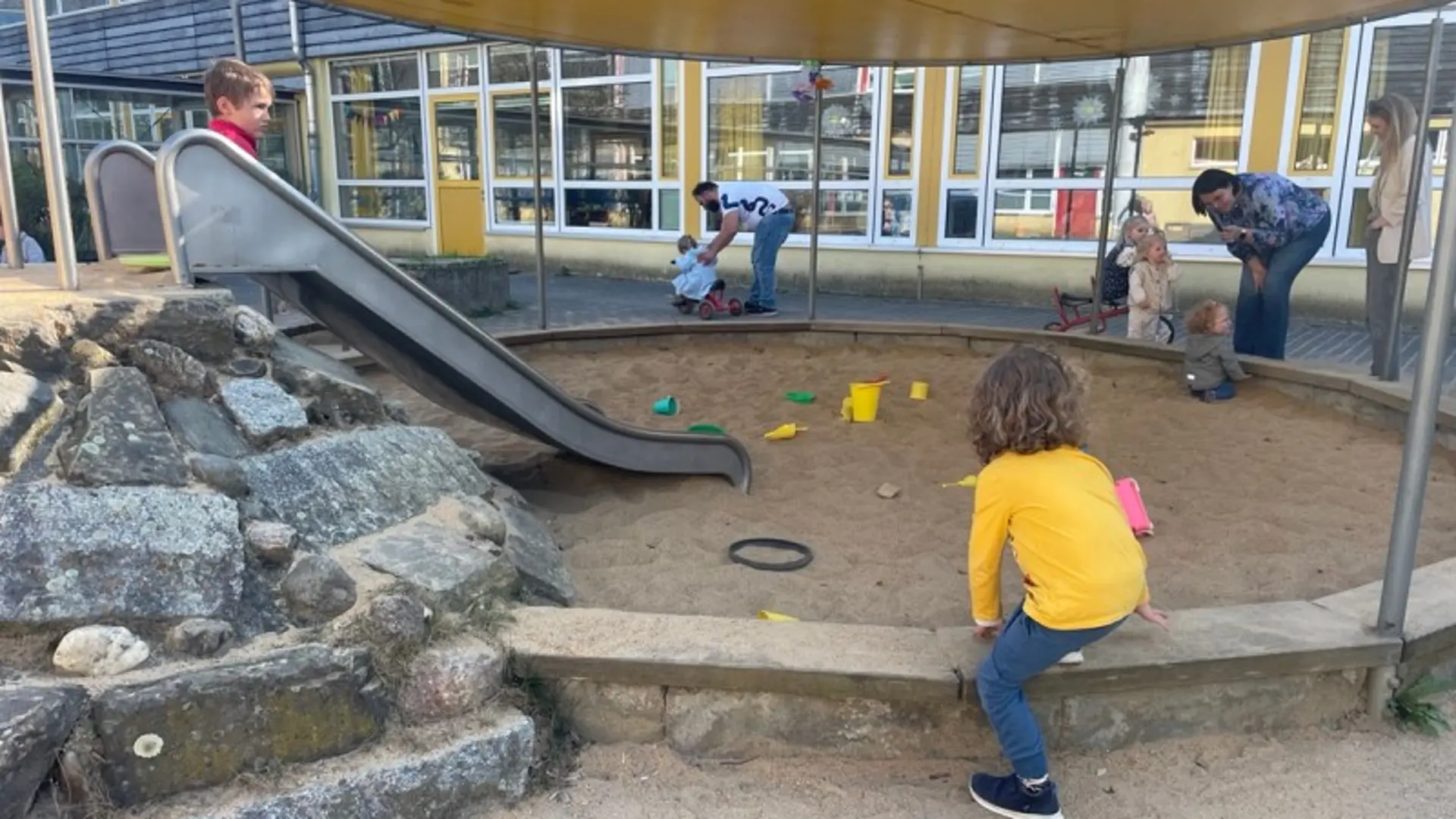 Kleine Kinder spielen in einem runden Sandkasten in den man auch über eine kleine Rutsche hineinrutsche kann