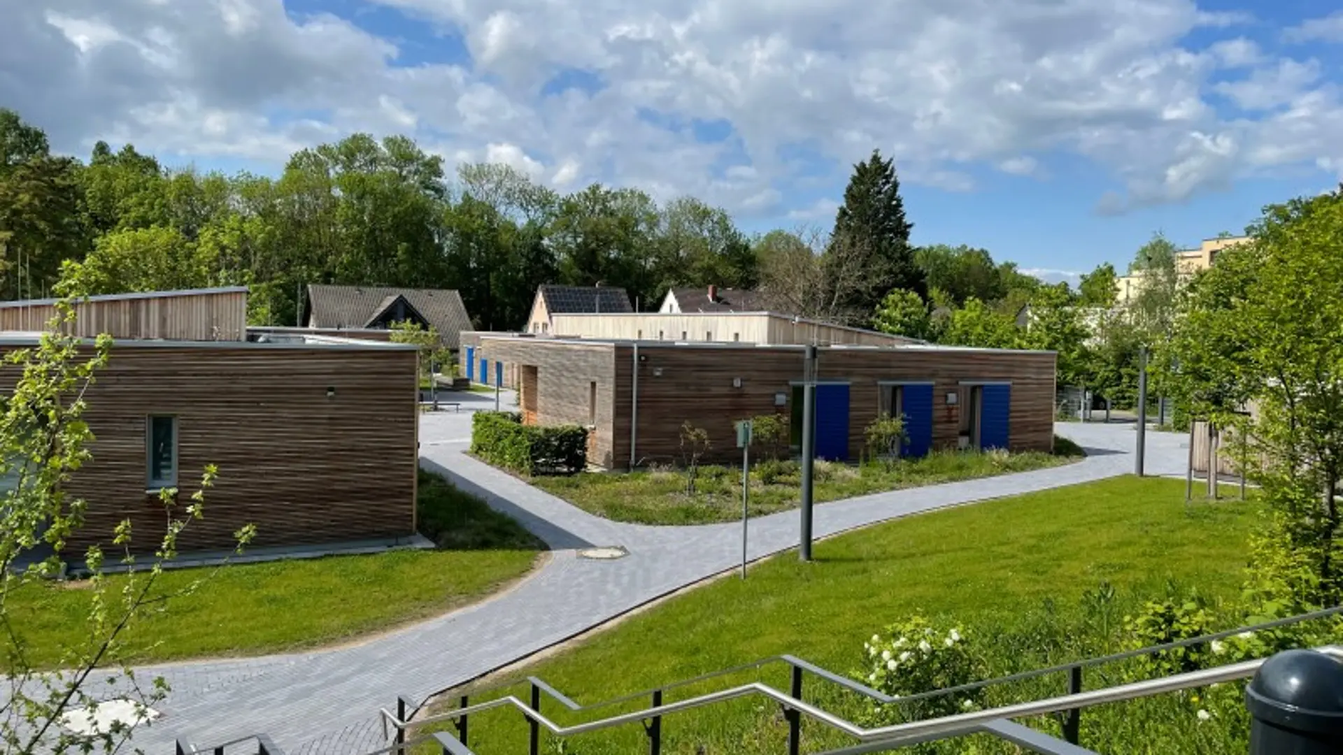 Moderne Holzgebäude mit blauen Akzenten, gepflasterte Wege, grüne Wiesen und eine Treppe unter bewölktem Himmel.