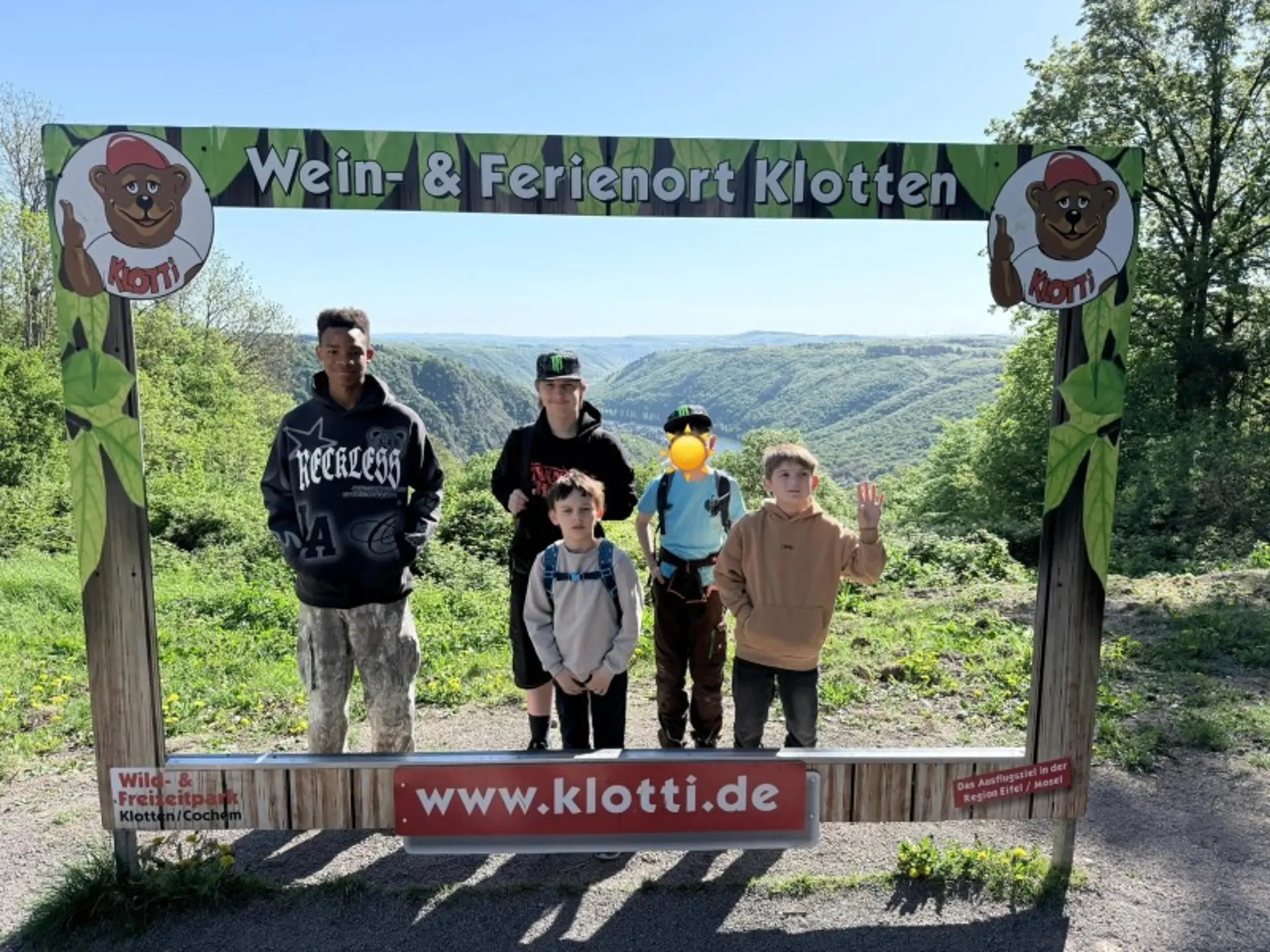 Fünf Kinder stehen in einem Holzrahmen des Wein- und Ferienorts Klotten. Hinter Ihnen ist das bewaldete Moseltal zu sehen.