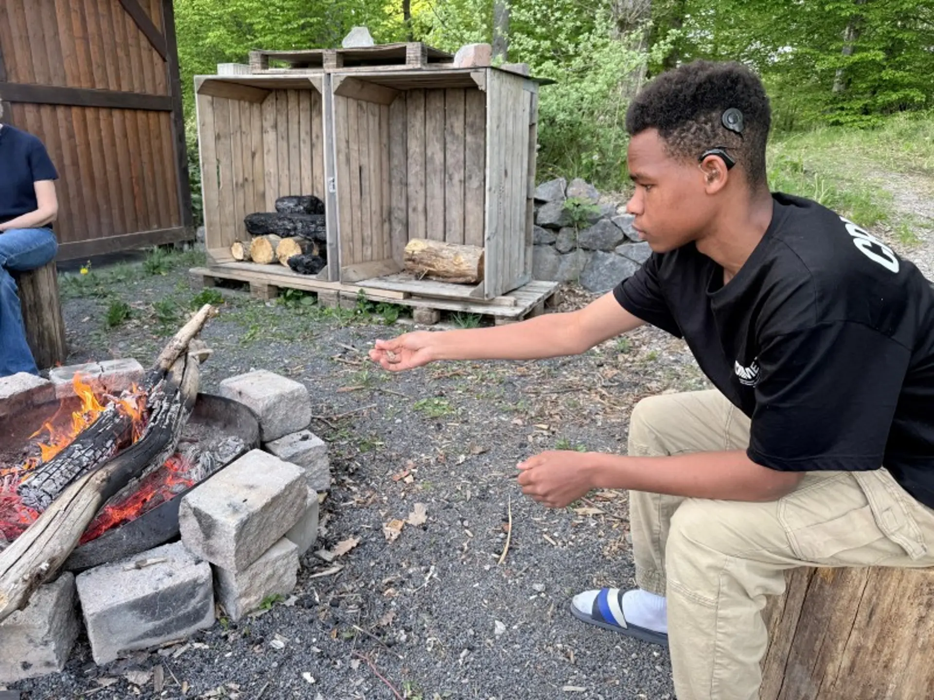 Ein Jugendlicher mit einem Cochlea-Implantat sitzt auf einem Sitz aus einem Baumstamm an einem Lagerfeuer neben einer Waldhütte.