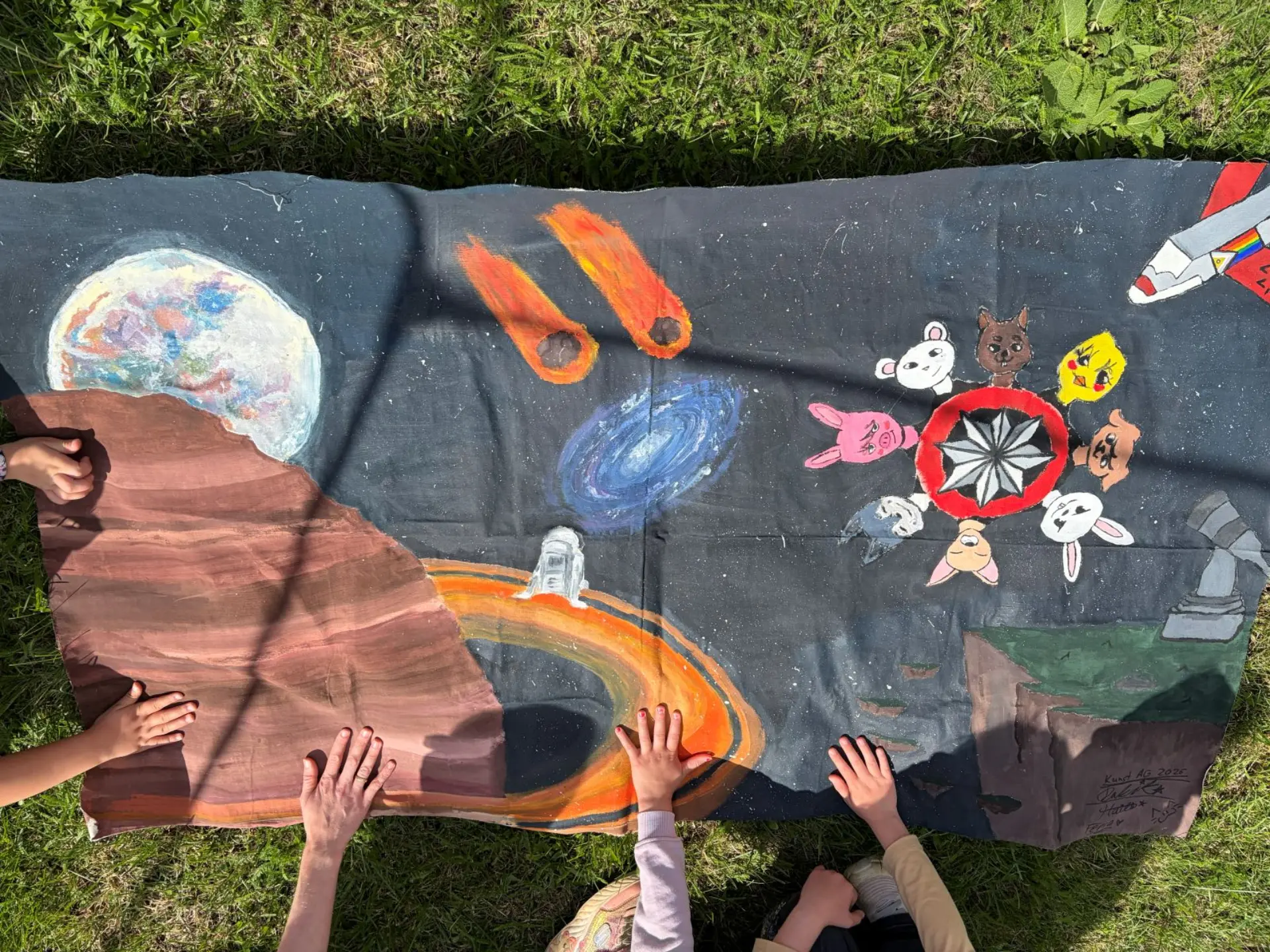 Auf einer Wiese liegt eine Stück einer Papierrolle, das mit Weltraum-Motiven (Milchstraße, Raumschiff, Meteoriten, ...) mit Plakafarben bunt bemalt ist