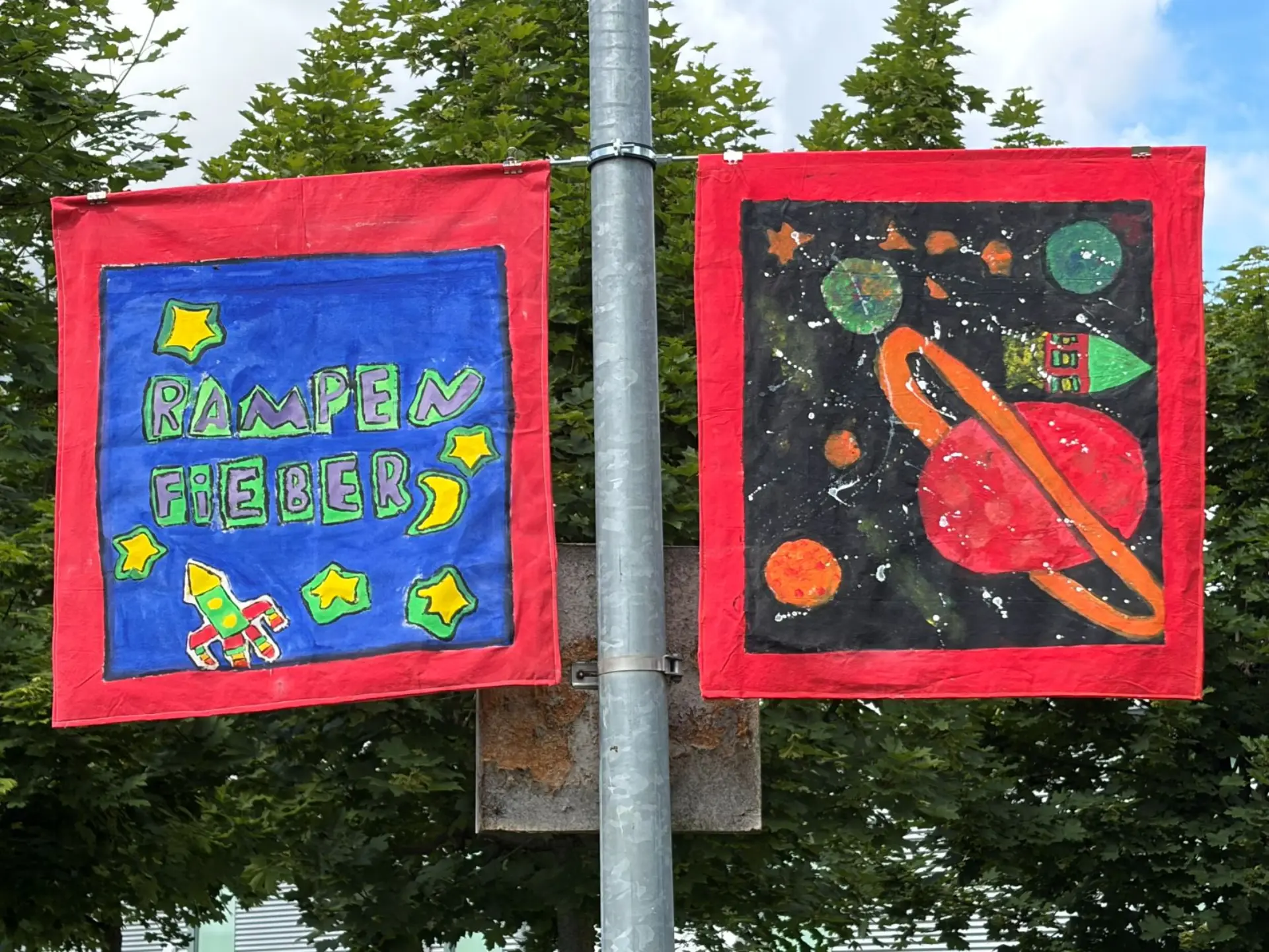Zwei quadratische Stoffbahnen sind an einem Straßenschild befestigt. Sie werben mit gemalten Weltraummotiven und dem Schriftzug Rampenfieber für das Kulturfestival.