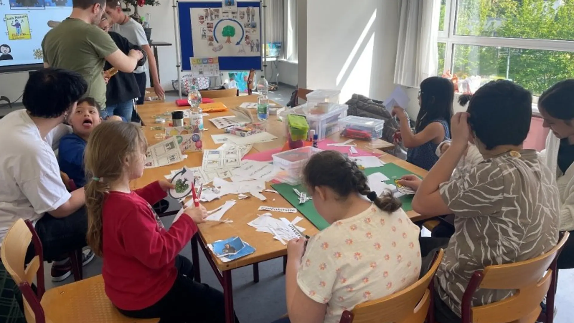 Mehrere Kinder und Erwachsene sitzen um einen kleinen Tisch und basteln und schneiden mit Papier