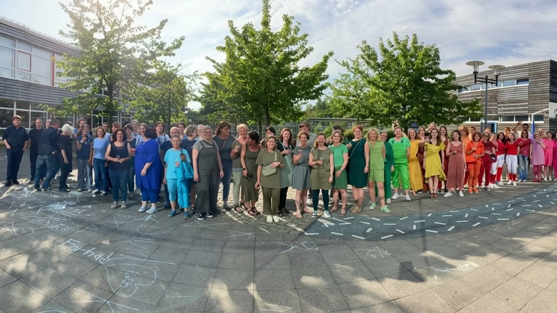 Gruppenbild der Lehrkräfte auf dem Schulhof. Sie sind in den Farben des Regenbogens angezogen: von links: lila, blau, grün, gelb, orange, rot