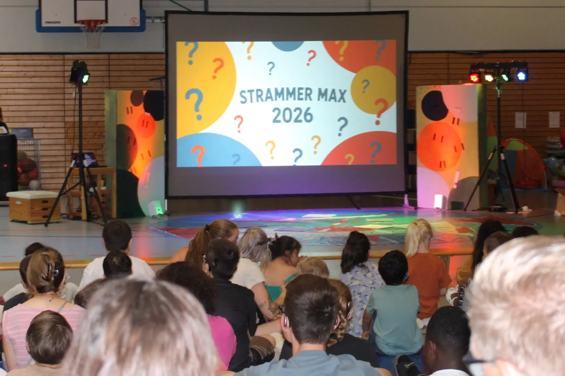 bunte Leinwand mit Aufschrift Strammer Max 2026 ??? davor sitzen Kinder und Erwachsene