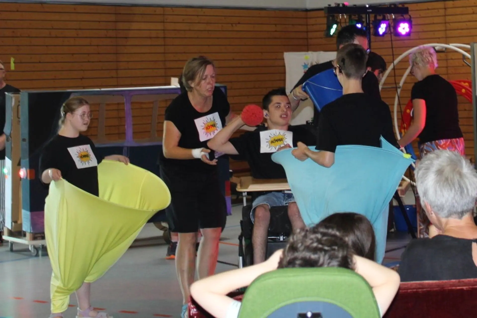 Junge und Mädchen stehen jeweils in einem Reifen mit blauen oder gelben Stoff. Jungendlicher im Rollstuhl legt Ball rein.