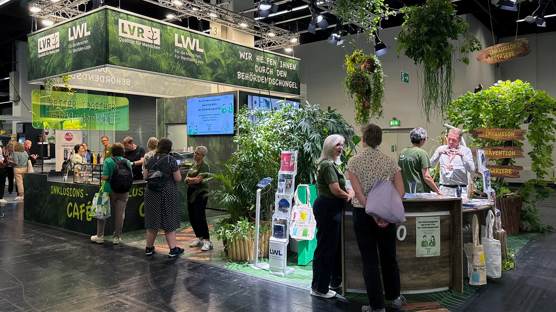 Messestand des Landschaftsverband Rheinland in grünen Farben mit Pflanzen, Empfangstresen, Bildschirmen sowie Besucherinnen und Besuchern in einer Messehalle.