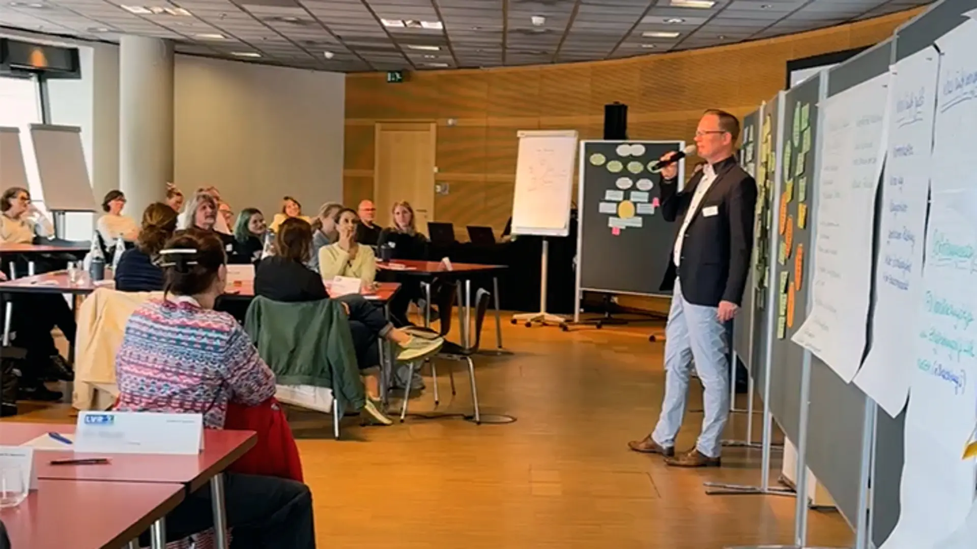 Ein Referent präsentiert Arbeitsergebnisse auf Moderationswänden vor einer Gruppe von Teilnehmenden in einem Workshop.
