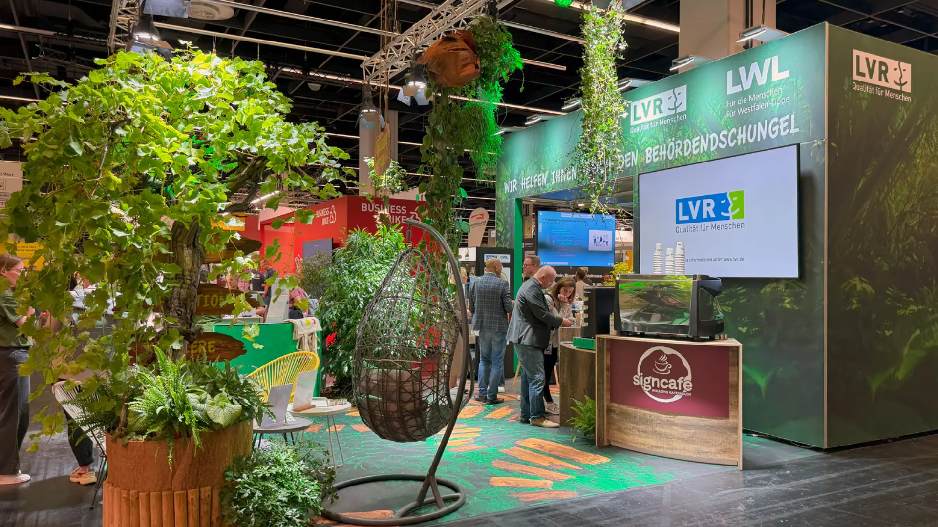 Messestand des Landschaftsverband Rheinland in grünen Farben mit Pflanzen, Empfangstresen, Bildschirmen sowie Besucherinnen und Besuchern in einer Messehalle.