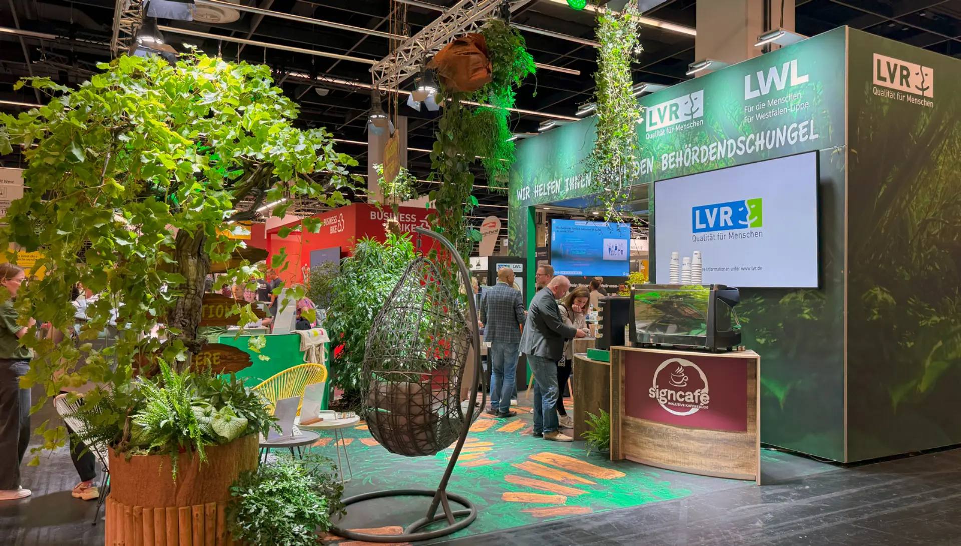 Seitlicher Blick auf den Messestand mit vielen Pflanzen, einer Theke des Sign Cafés und Sitzgelegenheiten.