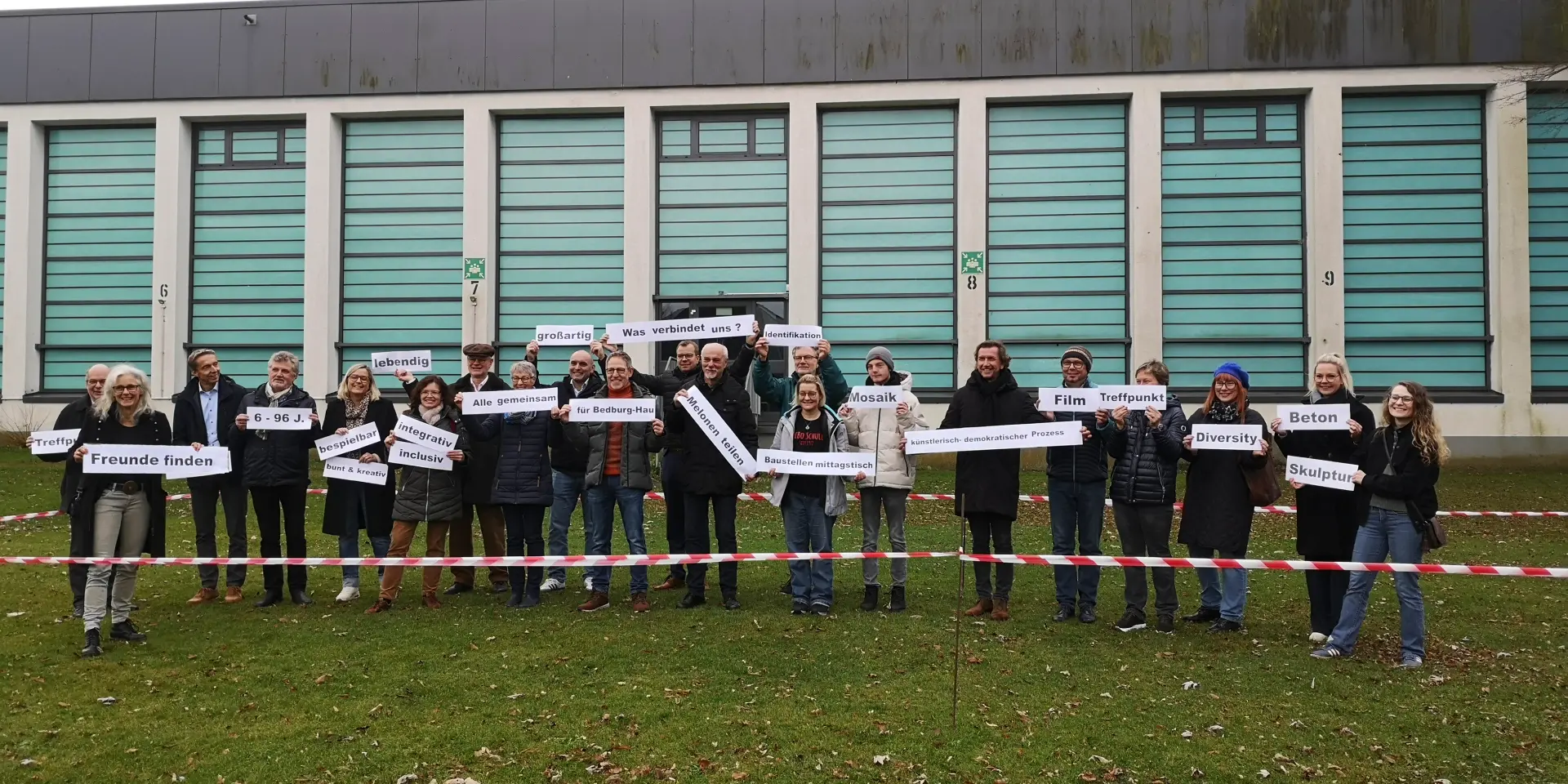 Eine groé Gruppe von Menschzen steht nebeneinander. Jeder Person hält ein großes Papier mit je einem Wort drauf in den Händen. Die Texte auf den Papieren: Treffpunkt, Freunde finden, 6-96 J., bespielbar, bunt & kreativ, lebendig, integrativ, inclusiv, alle gemeinsam, für Bedburg-Hau, Was verbindet uns?, großartig, Identifikation, Mosaik, Baustellenmittagstisch, Melonen teilen, künstlerisch-demokratischer Prozess, Film, Treffpunkt, Diversity, Beton, Skulptur 