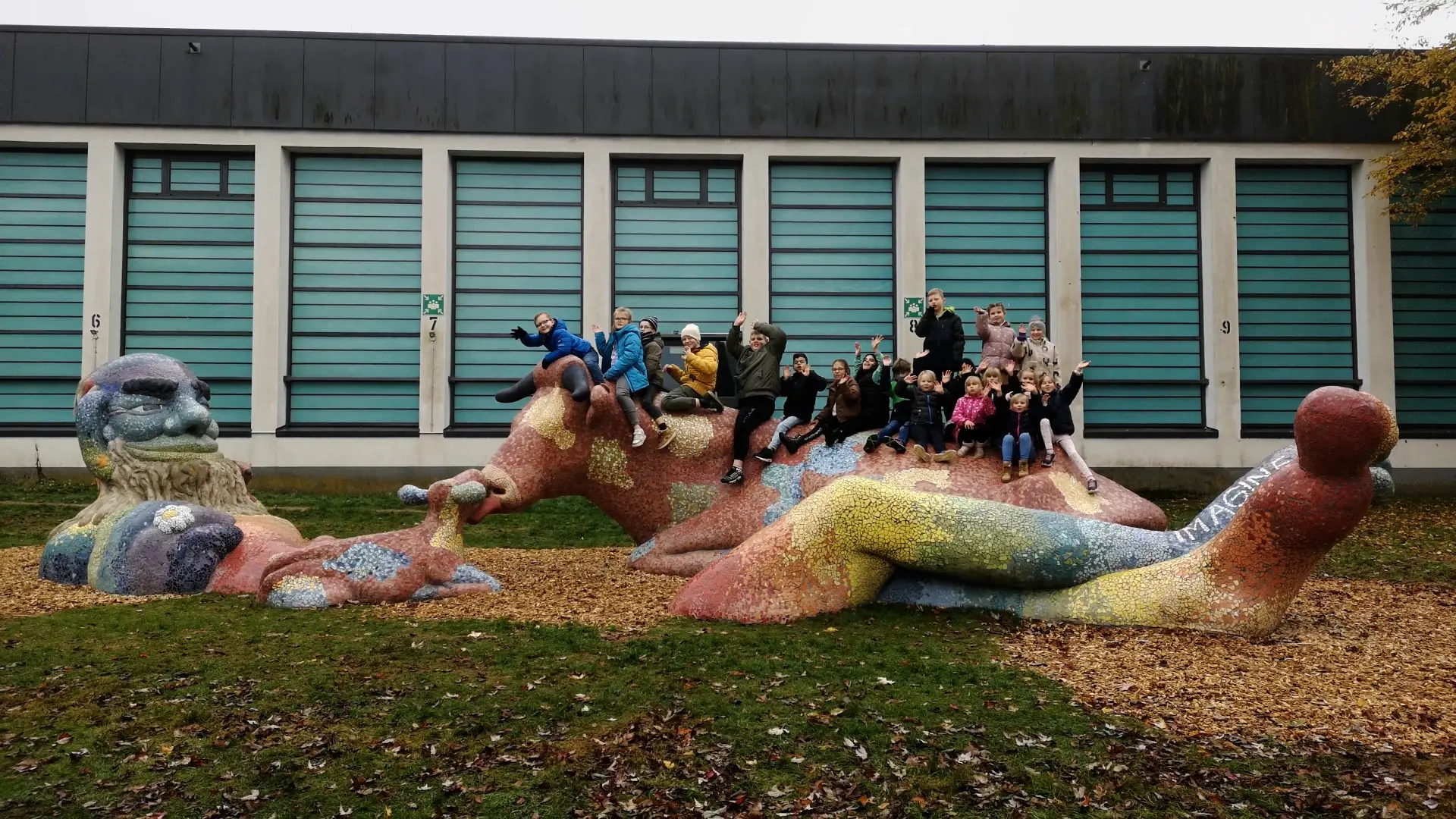 Eine Gruppe von Schülerinnen und Schülern sitzt auf einer Großskulptur und winkt zu.