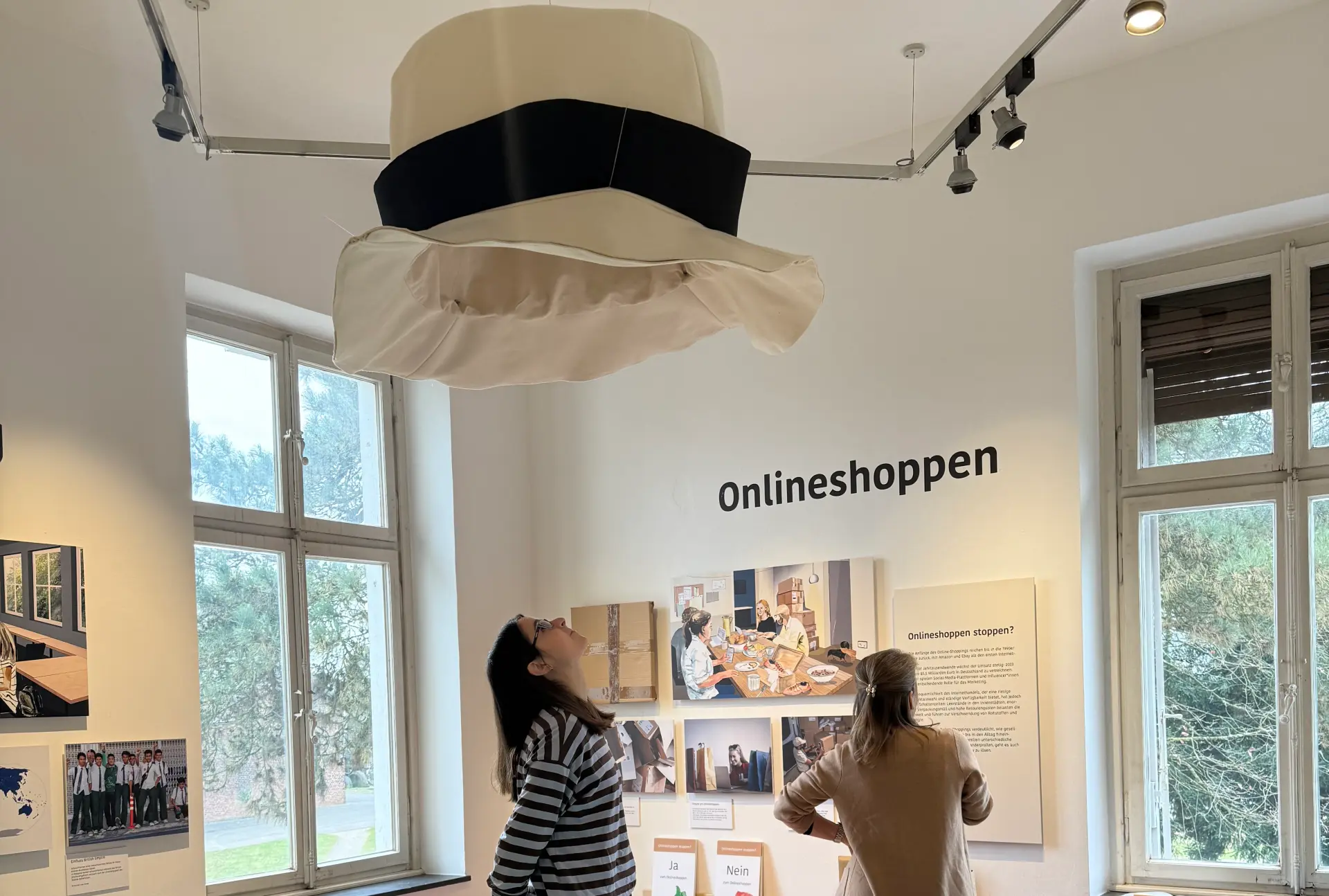 Zwei Frauen stehen in einem Ausstellungsraum. Eine Frau sieht sich einen Hut, der an der Decke hängt, an. Die zweite Frau liest einen Ausstellungstext.