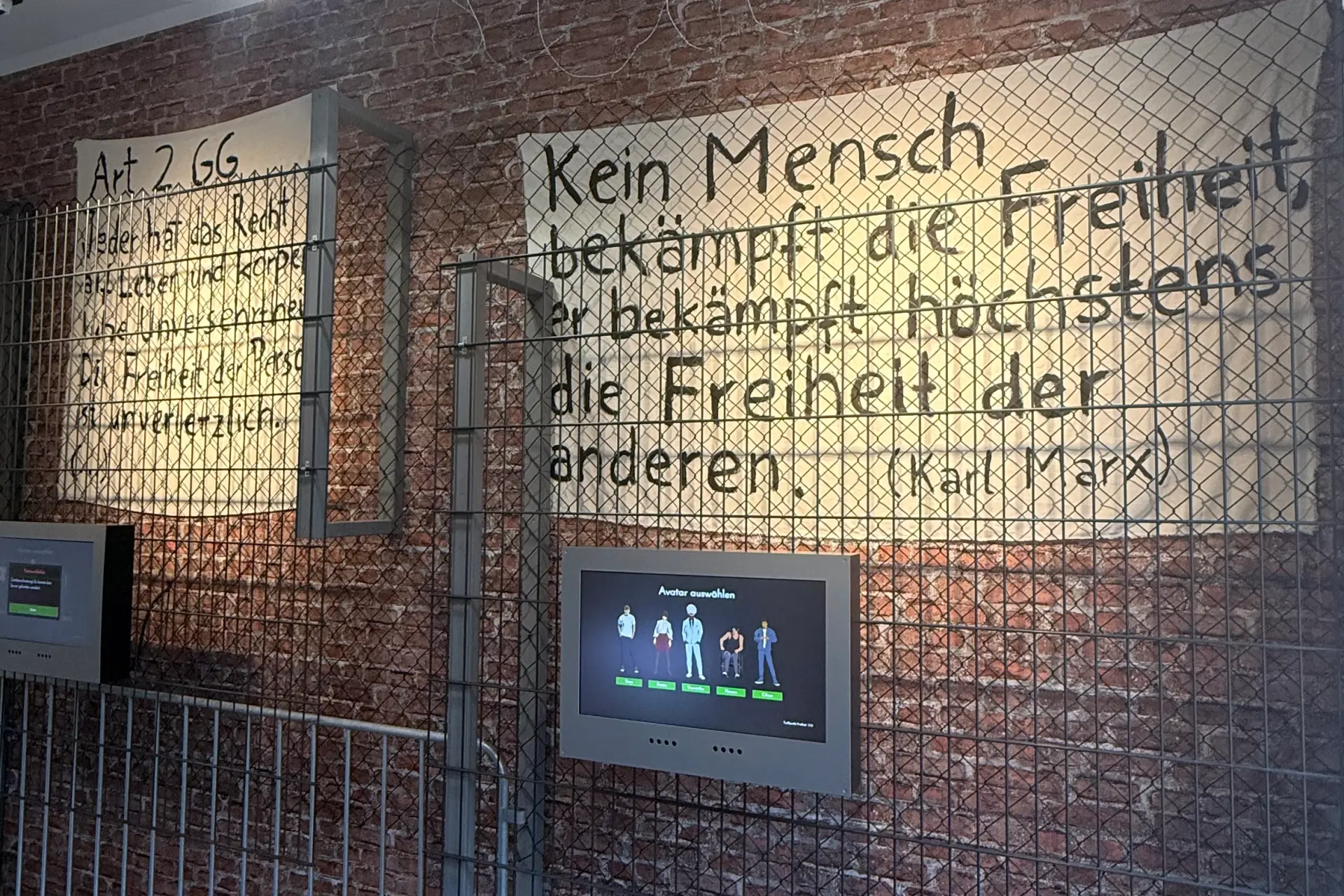 Eine Ziegelwand mit zwei Plakaten. Davor ein Absperrgitter. Text auf Plakat 1: Art. 2 GG. Jeder hat das Recht auf Leben und körperliche Unversehrtheit. Die Freiheit der Person ist unverletzlich. Text Plakat 2: Kein Mensch bekämpft die Freiheit, er bekämpft höchstens die Freiheit der anderen. (Karl Marx)